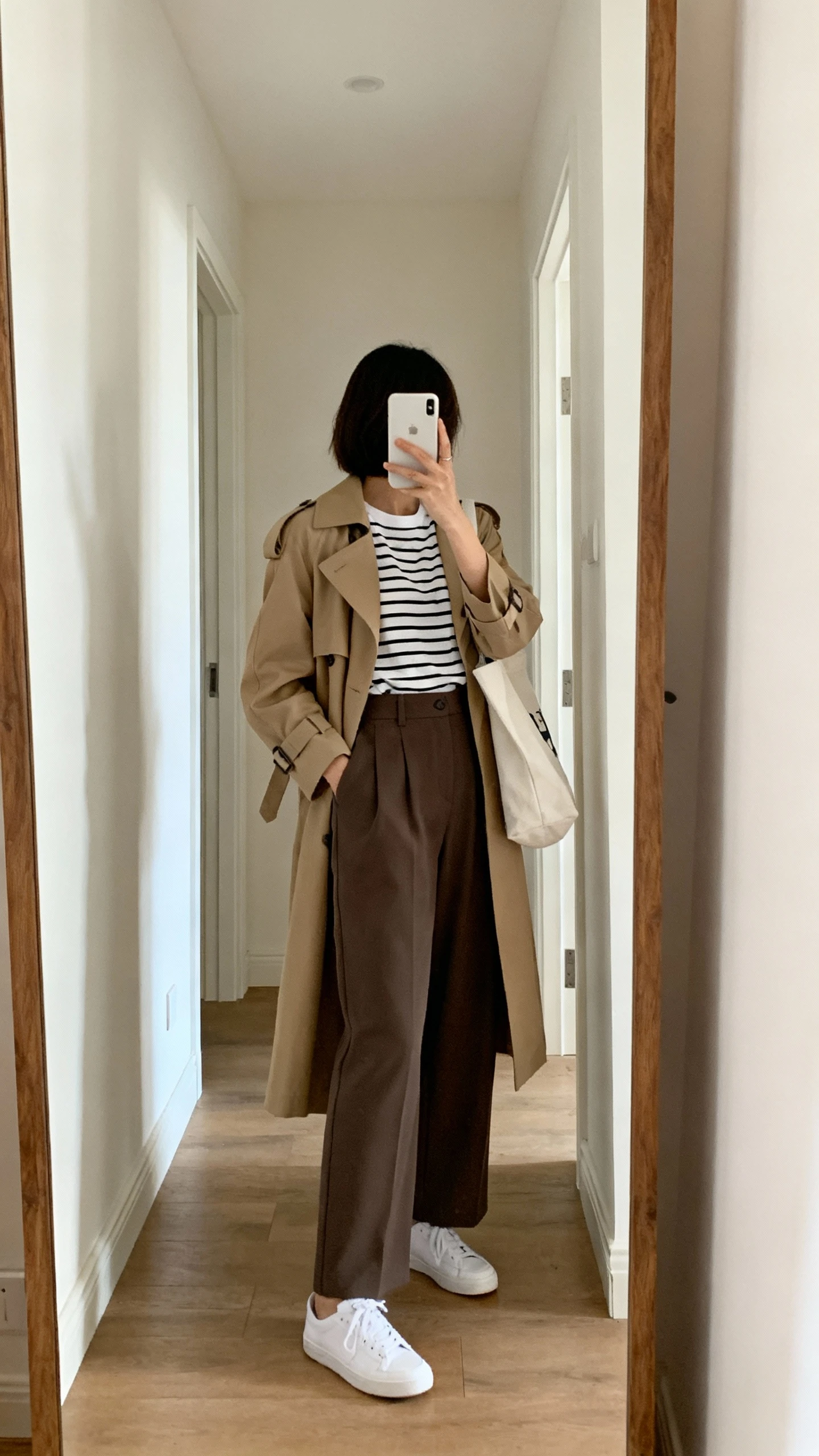 iPhone back-view hallway mirror shot of a woman wearing a classic trench coat over a Breton striped tee and high-waist wide-leg trousers with white sneakers and a tote bag, face not visible, natural daylight, casual iPhone photo.