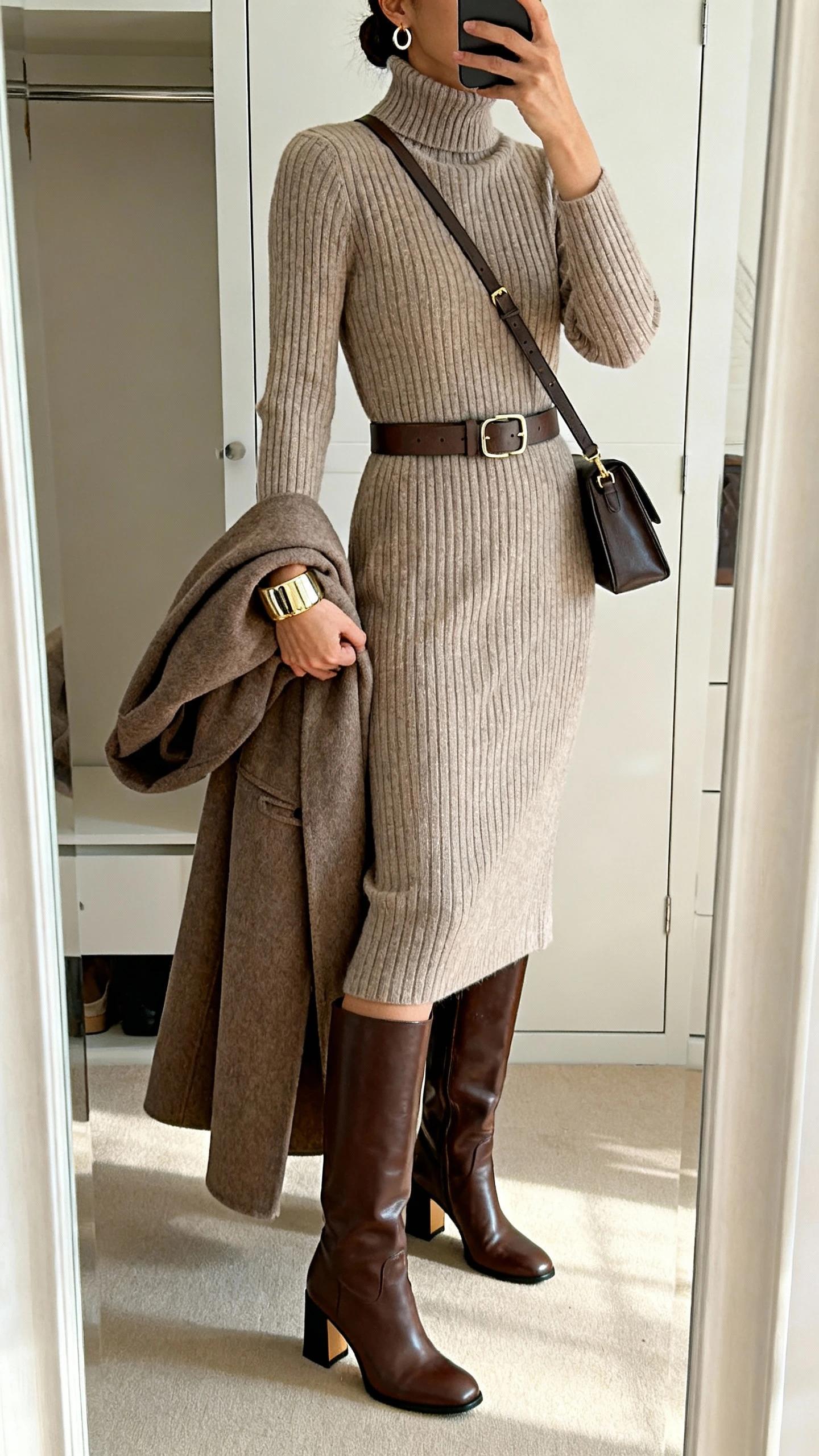 iPhone back-view mirror selfie of a woman wearing a ribbed knit midi dress (turtleneck), leather knee-high boots with a block heel, a waist belt, a longline wool coat over her arm, chunky cuff bracelet, small hoops, and a structured crossbody; face not visible, closet mirror, warm indoor lighting, iPhone photo quality.