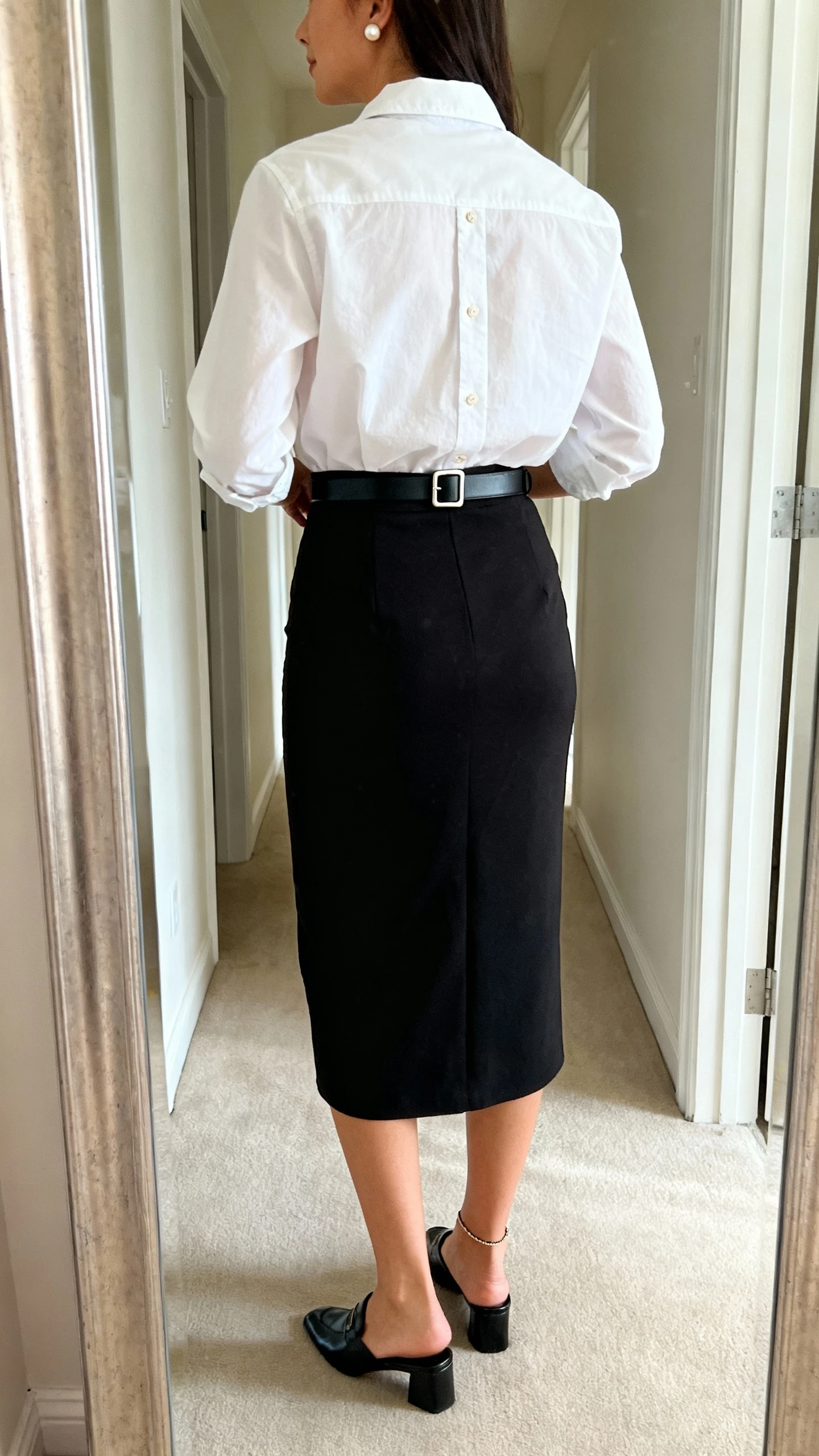 iPhone back-view mirror shot of a woman in a white cotton button-down shirt tucked into a high-waisted black midi pencil skirt, polished black mules, a delicate black belt with a clean buckle, and understated pearl studs, face not visible, hallway full-length mirror, indoor natural light, iPhone photo quality.