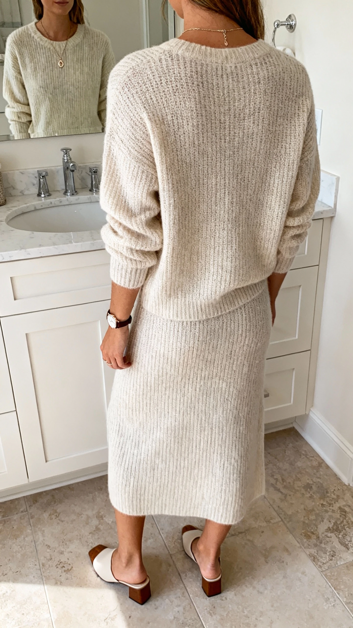 iPhone back view of a woman in an elevated matching knit set (fine-knit sweater and midi knit skirt), delicate pendant necklace, slim leather watch, low block-heel mules, face not visible, bathroom mirror, soft morning light, iPhone photo quality.