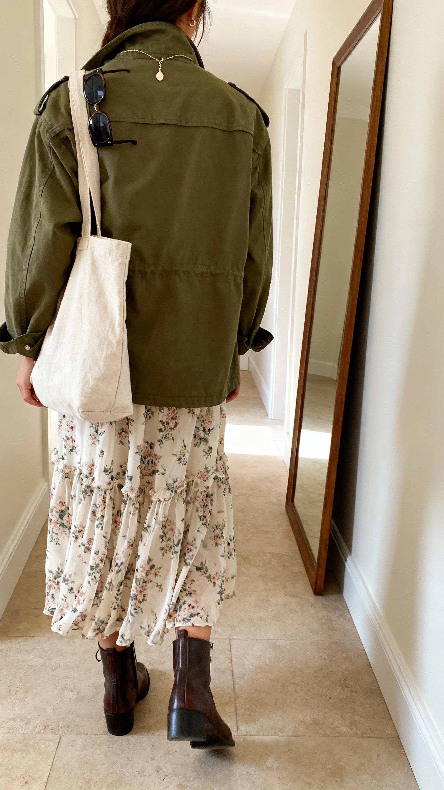 iPhone back view of a woman wearing a floaty floral midi dress layered with an olive utility jacket, ankle boots, canvas tote, sunglasses hooked on collar, delicate pendant, face not visible, hallway full-length mirror, natural daylight, iPhone photo quality.