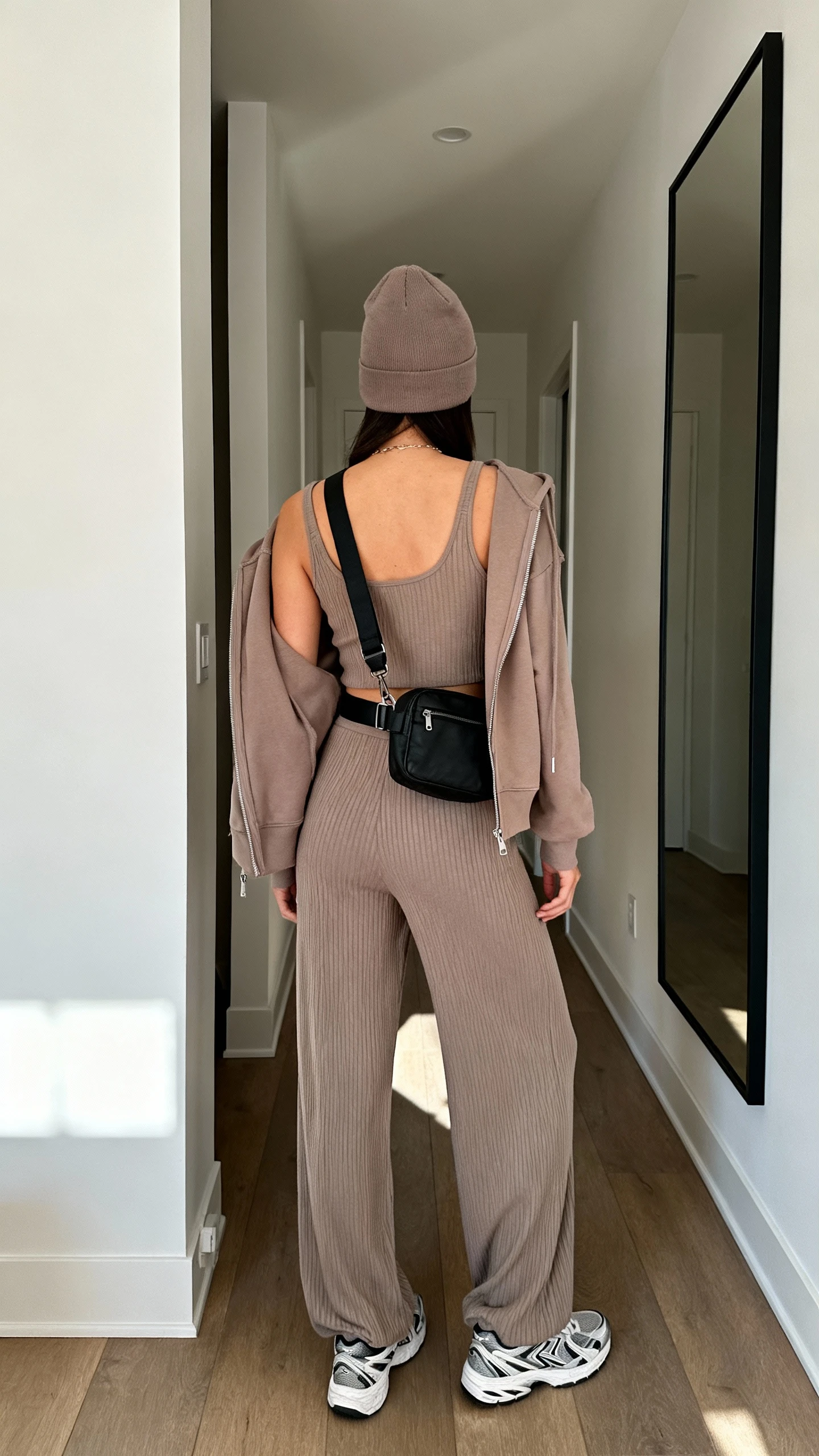iPhone back view of a woman wearing a matching ribbed knit set (taupe tank and wide-leg pants), a tonal zip-up hoodie, monochrome sneakers, a black belt bag worn crossbody, and a minimal beanie, face not visible, hallway mirror, natural daylight, casual iPhone photo.
