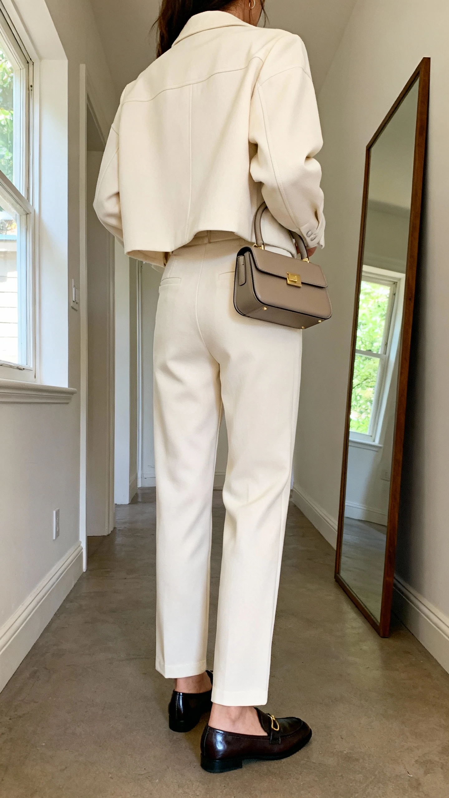 iPhone back view of a woman wearing a monochrome matching set (boxy jacket and straight-leg trousers) with sleek loafers and a structured top-handle bag, face not visible, hallway full-length mirror, natural daylight from window, iPhone photo quality.