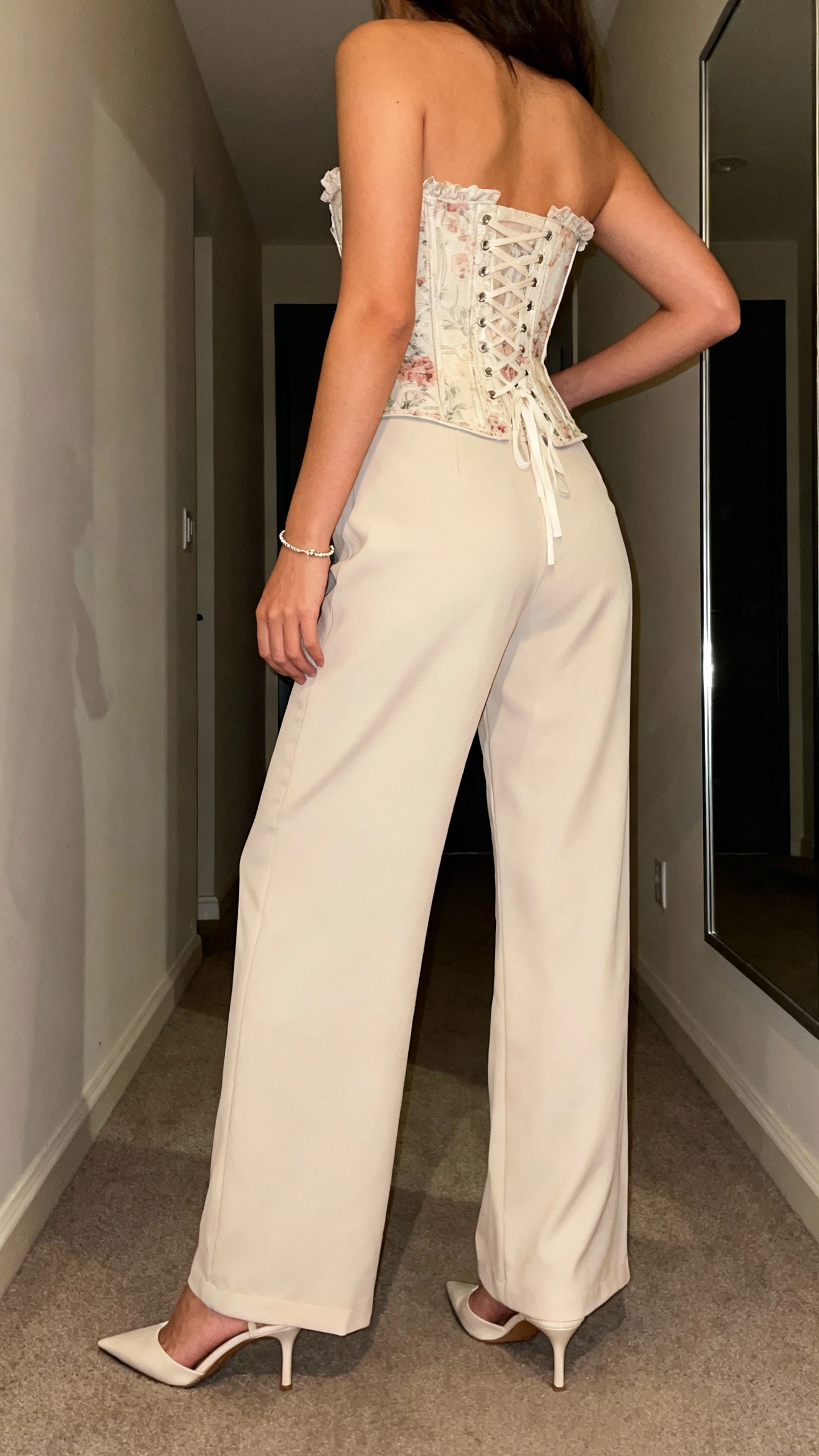 iPhone back view of a woman wearing a romantic corset top with wide-leg trousers, pointed-toe heels, and a dainty bracelet, face not visible, hallway mirror, evening indoor lighting, iPhone photo quality.