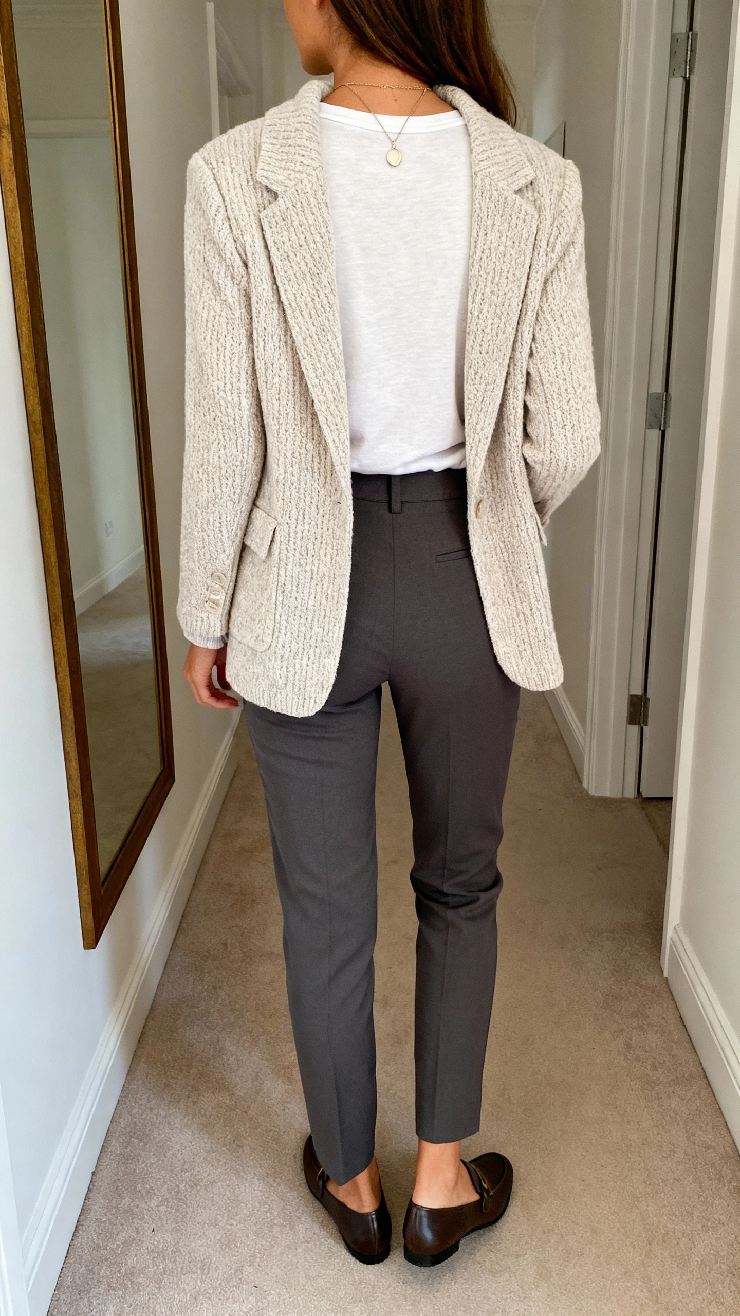 iPhone back view of a woman wearing a soft knit blazer over a fitted crewneck tee with stretch tailored pants and loafers, delicate pendant necklace, face not visible, hallway mirror, natural daylight, iPhone photo quality.