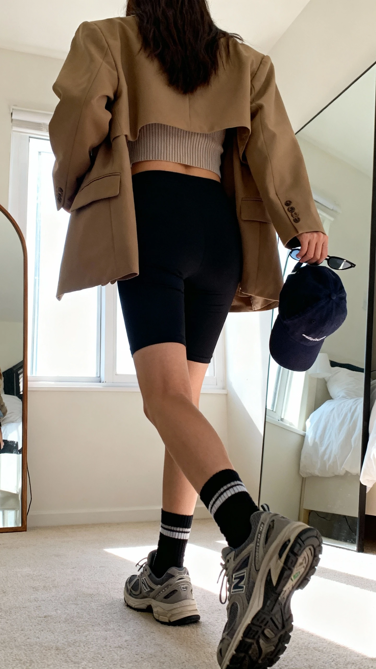 iPhone back view of a woman wearing black bike shorts, oversized tan blazer, ribbed crop tank, crew socks with chunky sneakers, baseball cap tucked in hand, and slim sunglasses, face not visible, casual walking pose, bedroom mirror, bright window daylight, iPhone photo quality.