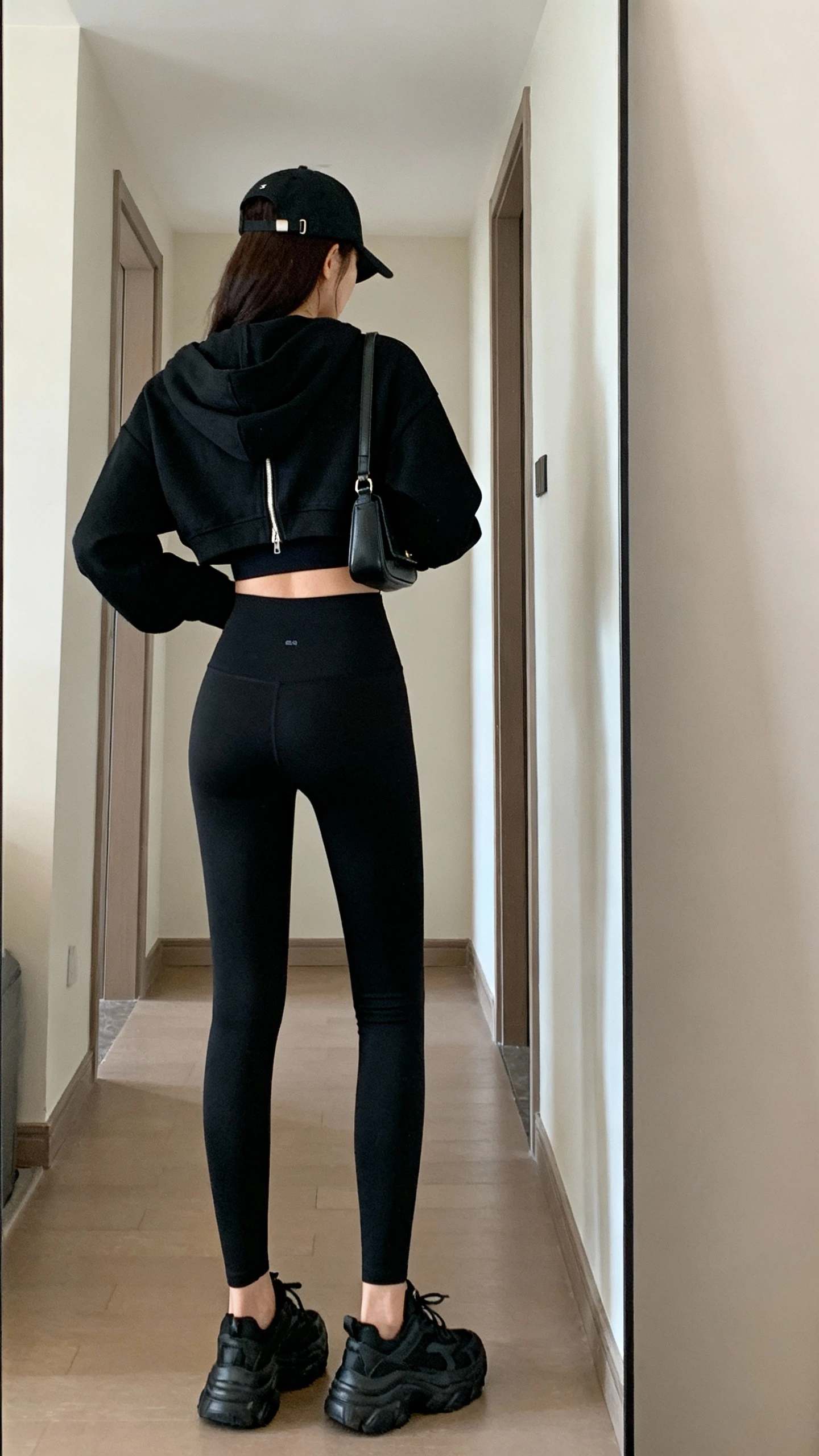 iPhone back view of a woman wearing model-off-duty monochrome athleisure: black zip-up hoodie with matching black sports bra, high-waist black leggings, chunky black trainers, black baseball cap and tiny shoulder bag, face not visible, hallway full-length mirror, soft indoor lighting, iPhone photo quality.
