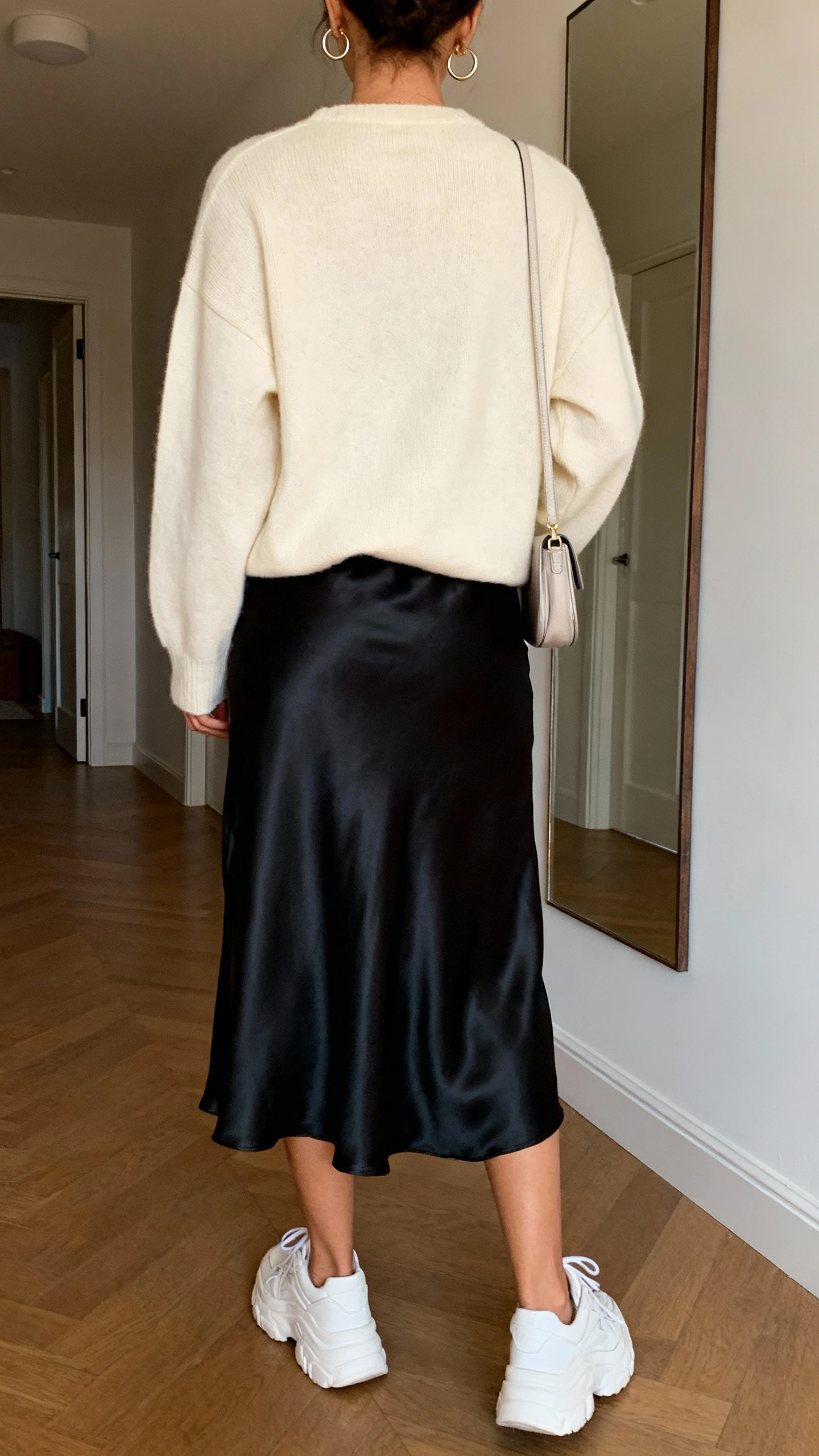 iPhone back-view photo of a woman in a black satin slip skirt with a soft cream crewneck sweater and chunky white sneakers, dainty hoop earrings, small shoulder bag, face not visible, hallway mirror, warm indoor lighting, iPhone photo quality.