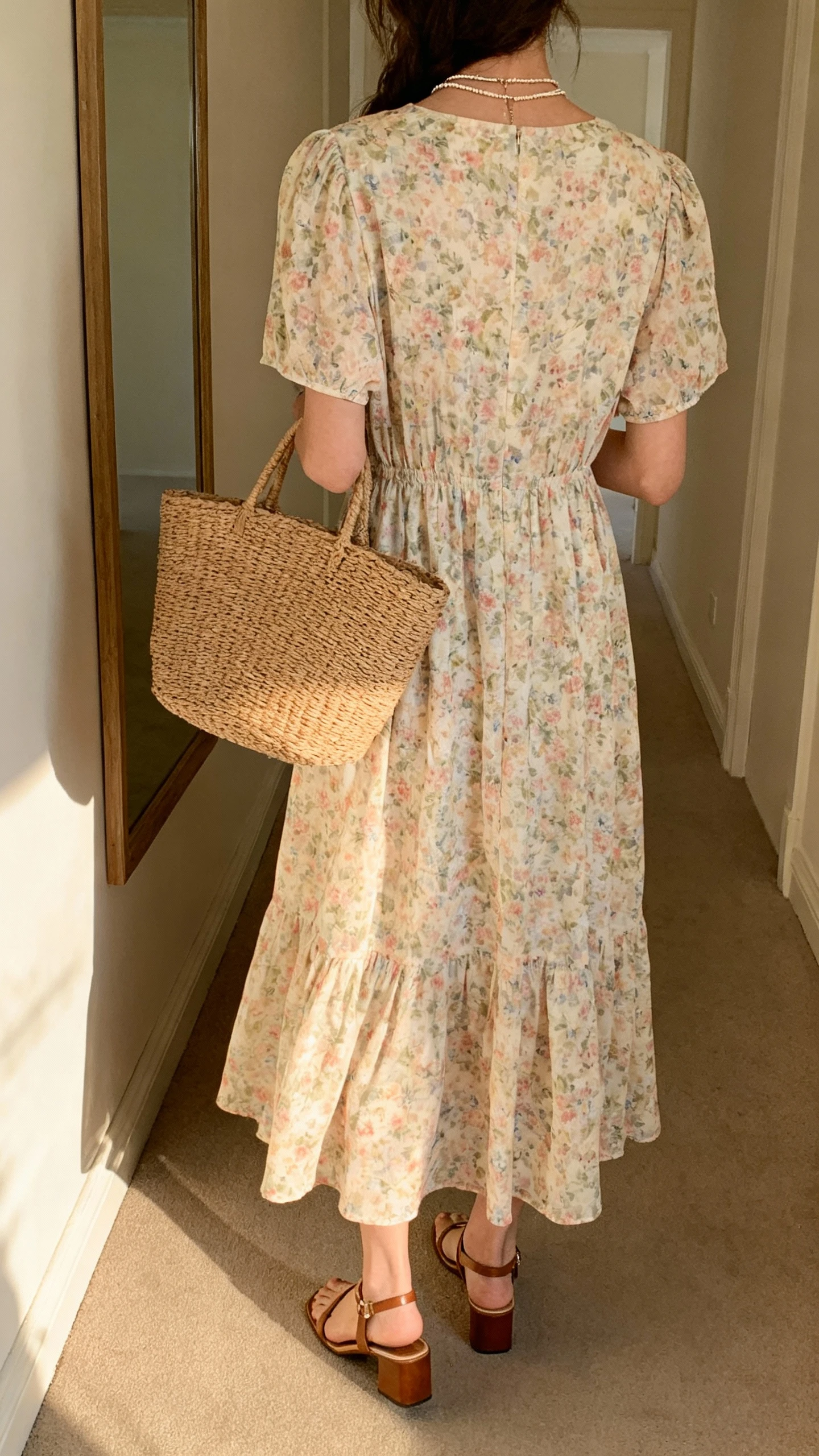iPhone back-view photo of a woman wearing a breezy floral midi dress with short sleeves, low block-heel sandals, a woven straw tote, and dainty layered necklaces, face not visible, hallway mirror with soft afternoon light, casual iPhone photo quality.