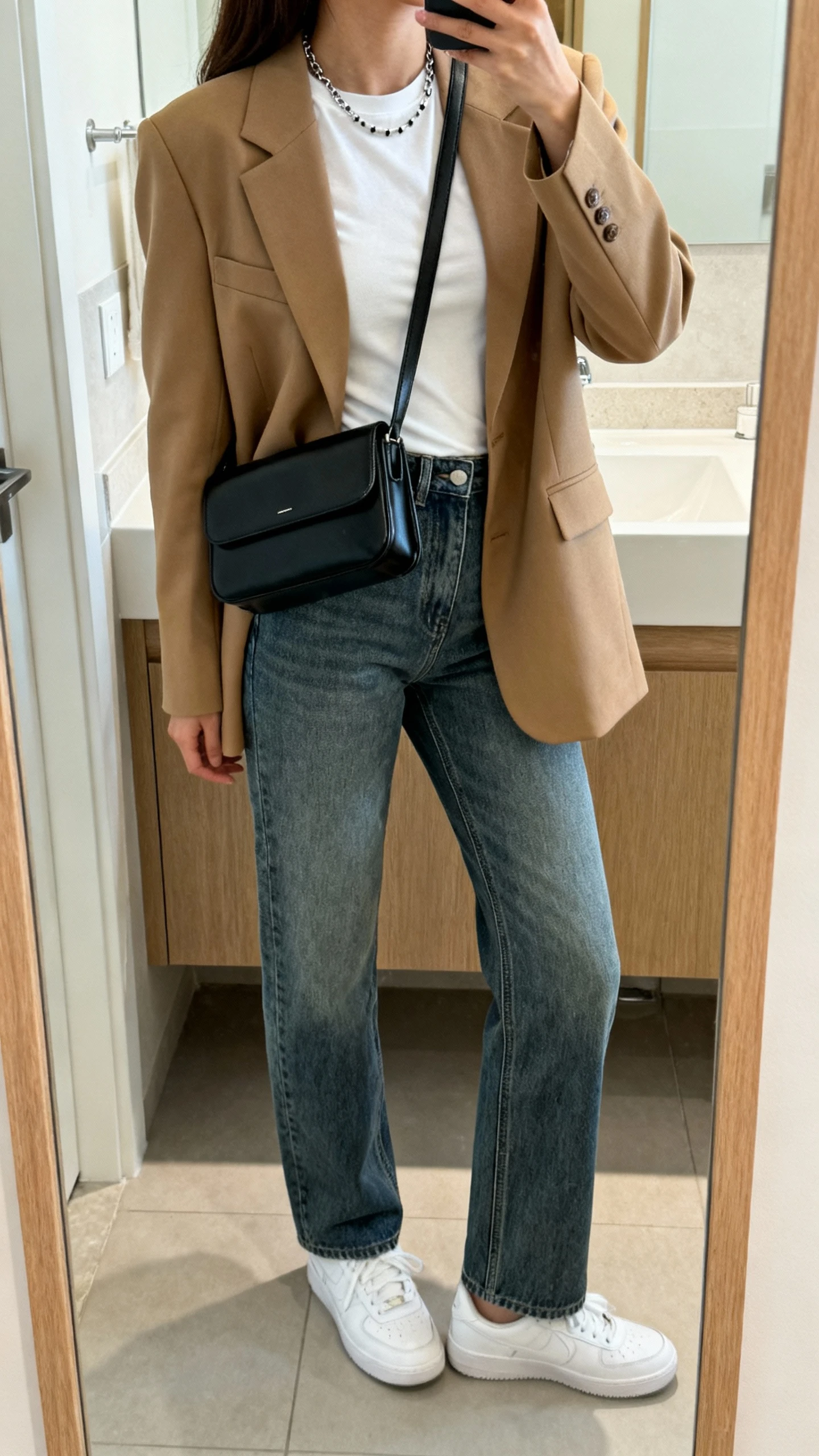 iPhone back-view selfie of a woman wearing mid-rise straight-leg medium-wash jeans, a camel tailored blazer, fitted white tee, white sneakers, thin chain necklace with subtle studs, and a sleek black crossbody bag, face not visible, bathroom mirror, natural daylight, iPhone photo quality.