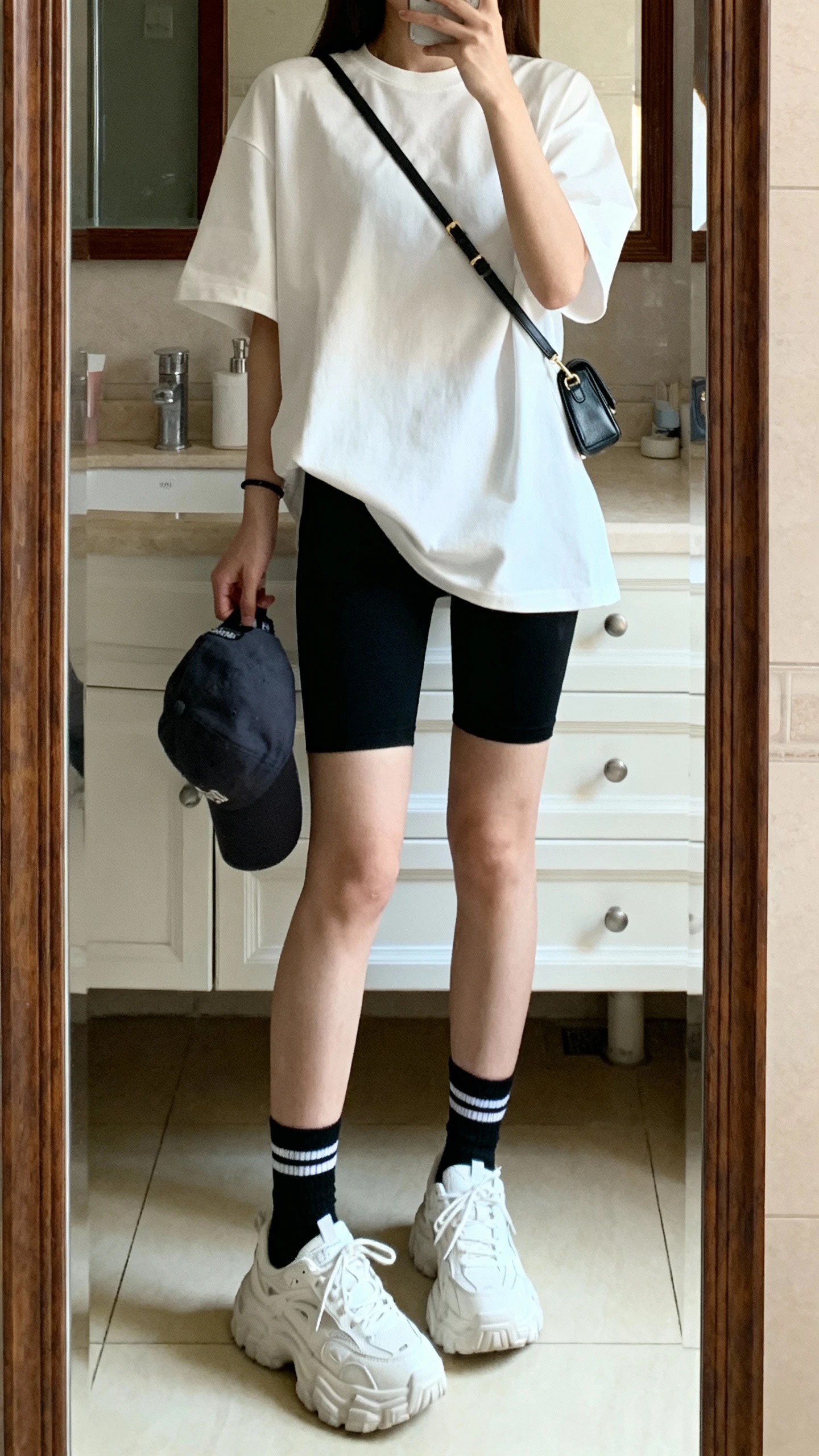 iPhone full-length mirror selfie of a woman wearing a boxy white tee with black bike shorts, chunky crew socks and white trainers, baseball cap in hand and a small crossbody bag, face not visible, bathroom mirror, soft indoor morning light, iPhone photo quality.
