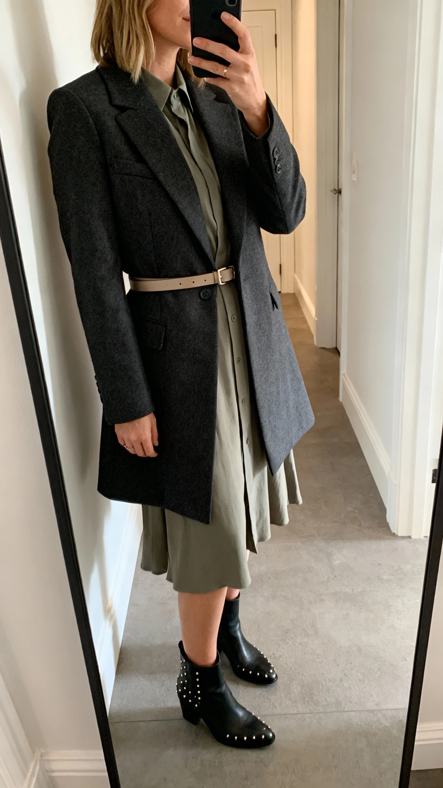 iPhone hallway mirror selfie of a woman wearing a smart-casual dress layered with a sharp tailored coat and a slim belt, ankle boots, subtle studs, face not visible, side angle, natural indoor lighting, iPhone photo quality.