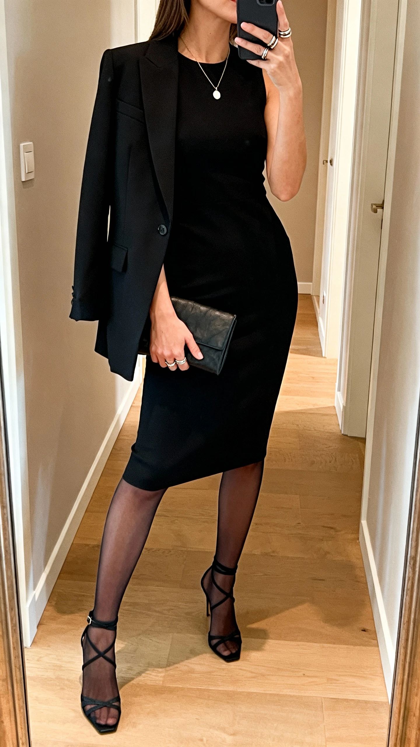 iPhone mirror selfie of a woman in a classic little black sheath dress with strappy heels, tailored black blazer draped over shoulders, delicate pendant necklace, stackable rings, small clutch, and sheer black tights, face not visible, side angle in a hallway mirror, warm indoor lighting, iPhone photo quality.
