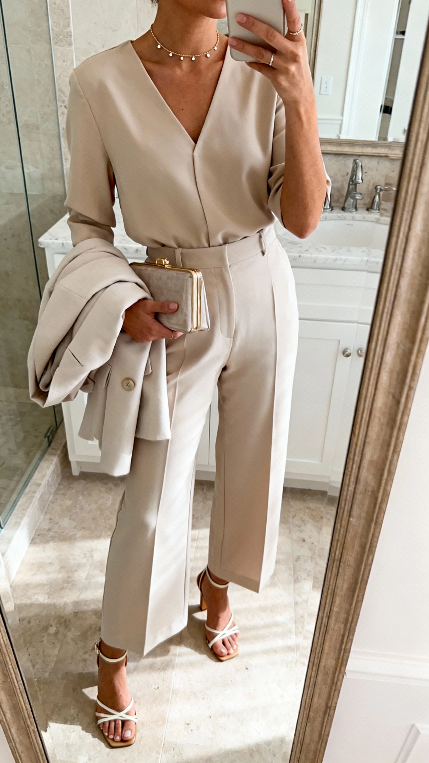 iPhone mirror selfie of a woman in a coordinated set (tailored top with wide-leg pants), strappy heeled sandals, a fine-chain choker with tiny stud earrings, and a compact clutch, with a light blazer folded over her forearm; face not visible, cropped at neck, bathroom mirror, soft morning light, iPhone photo quality.