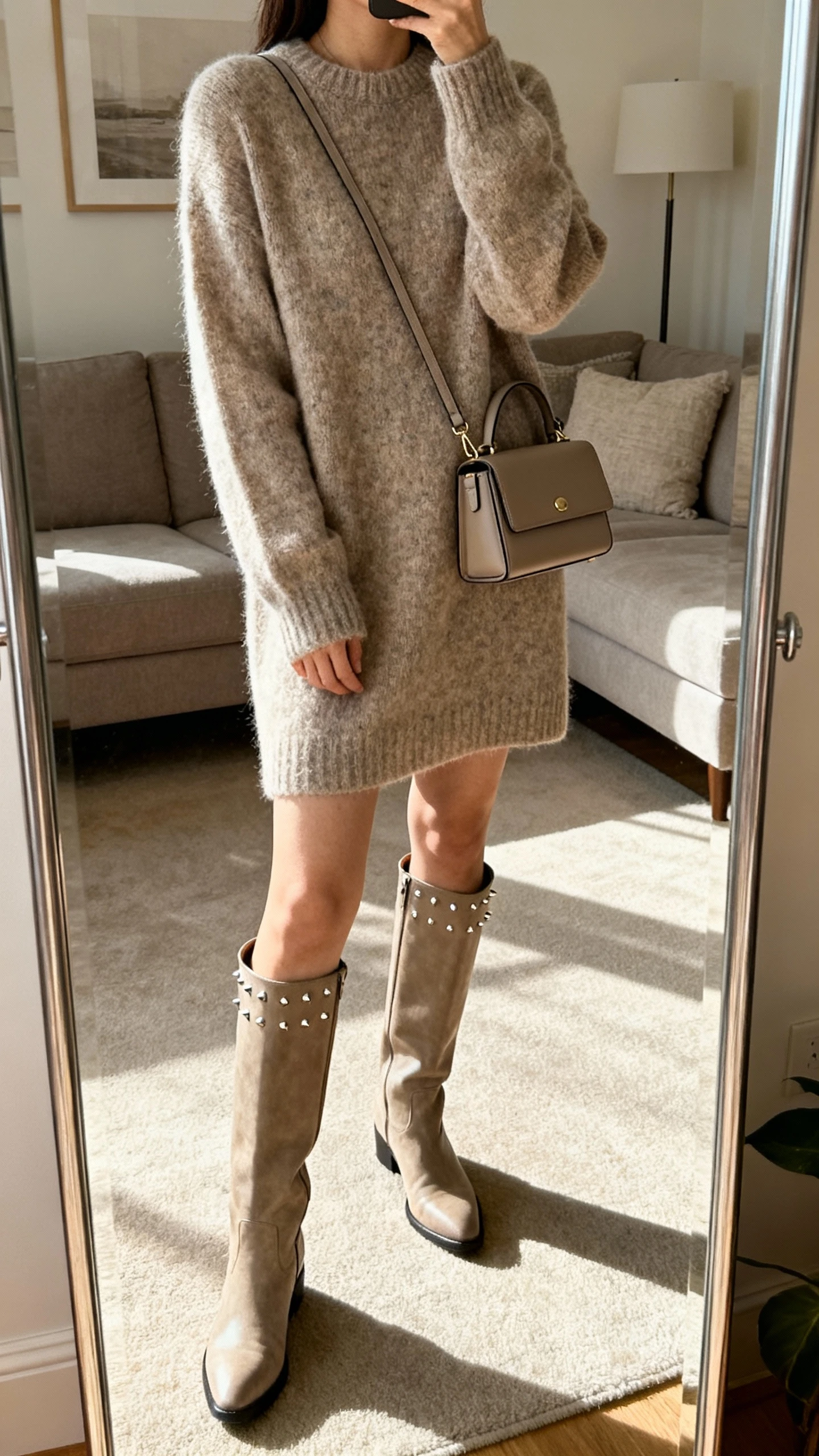 iPhone mirror selfie of a woman in a cozy sweater dress with tall boots, minimal studs, and a structured mini bag, face not visible, cropped at neck, living room full-length mirror, soft afternoon light, iPhone photo quality.