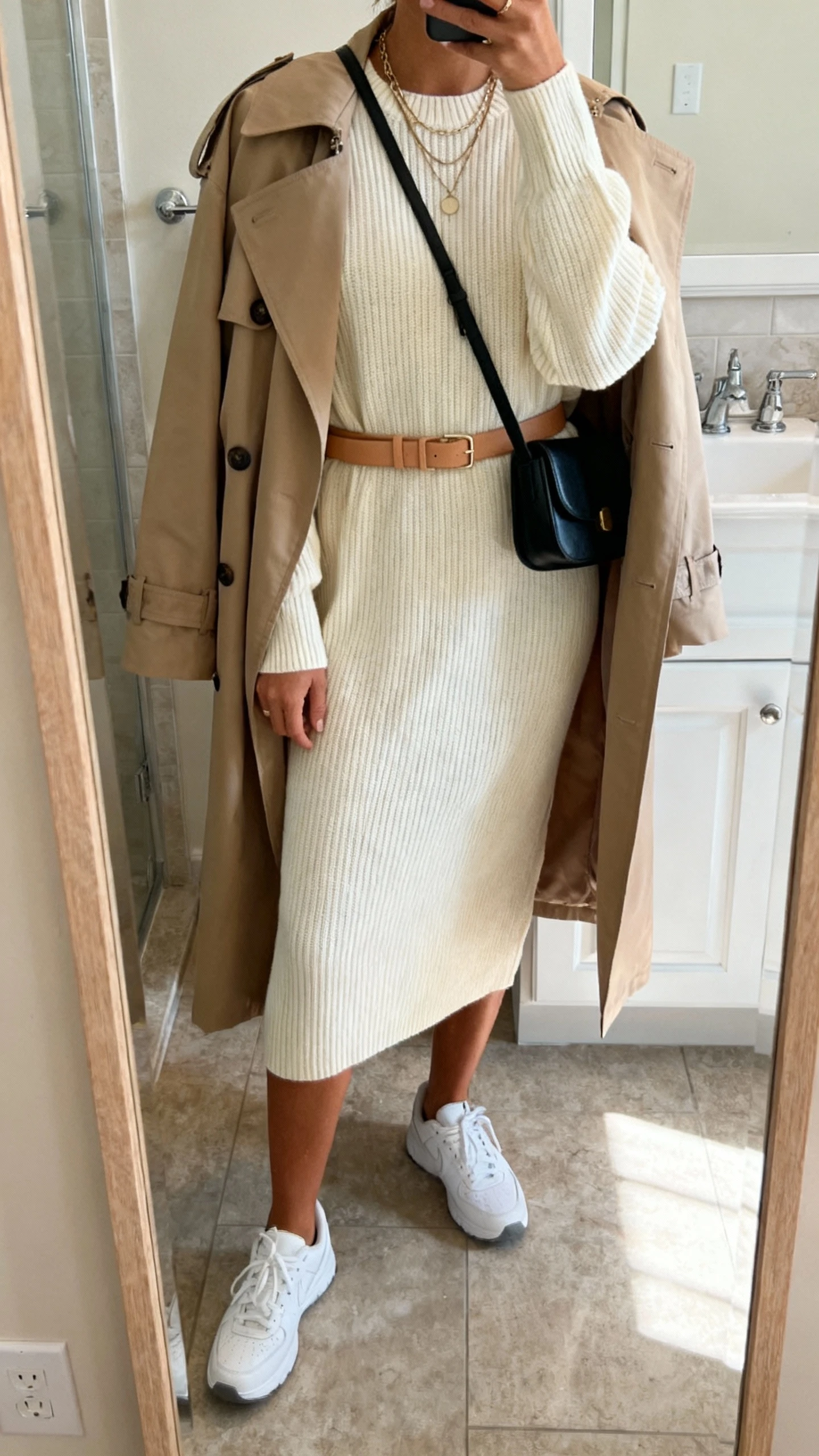 iPhone mirror selfie of a woman in a cream ribbed midi sweater dress with a slim tan belt, clean white sneakers, a beige trench coat draped over shoulders, a black crossbody bag, and a layered necklace, face not visible, bathroom full-length mirror, soft indoor light, iPhone photo quality.