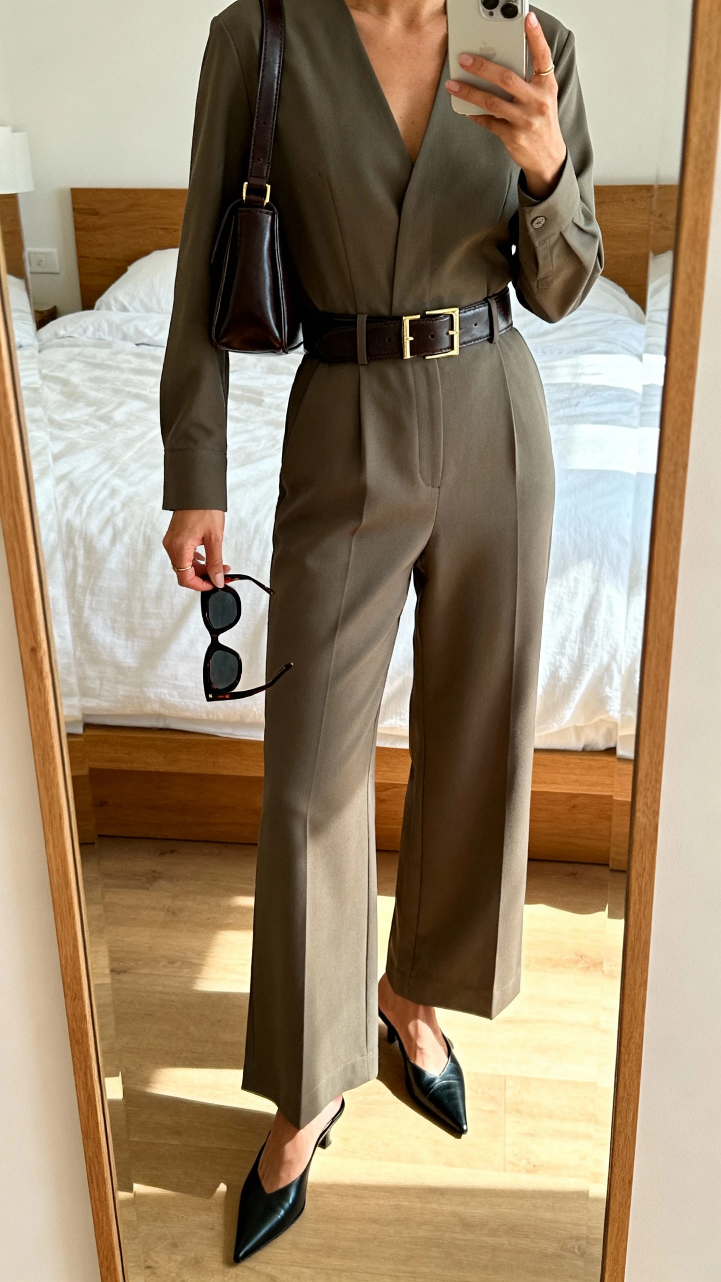 iPhone mirror selfie of a woman in a minimal tailored jumpsuit with a sharp leather belt, structured shoulder bag, pointed mules, and bold sunglasses in hand, face not visible, cropped at shoulders, bedroom mirror, natural daylight, iPhone photo quality.