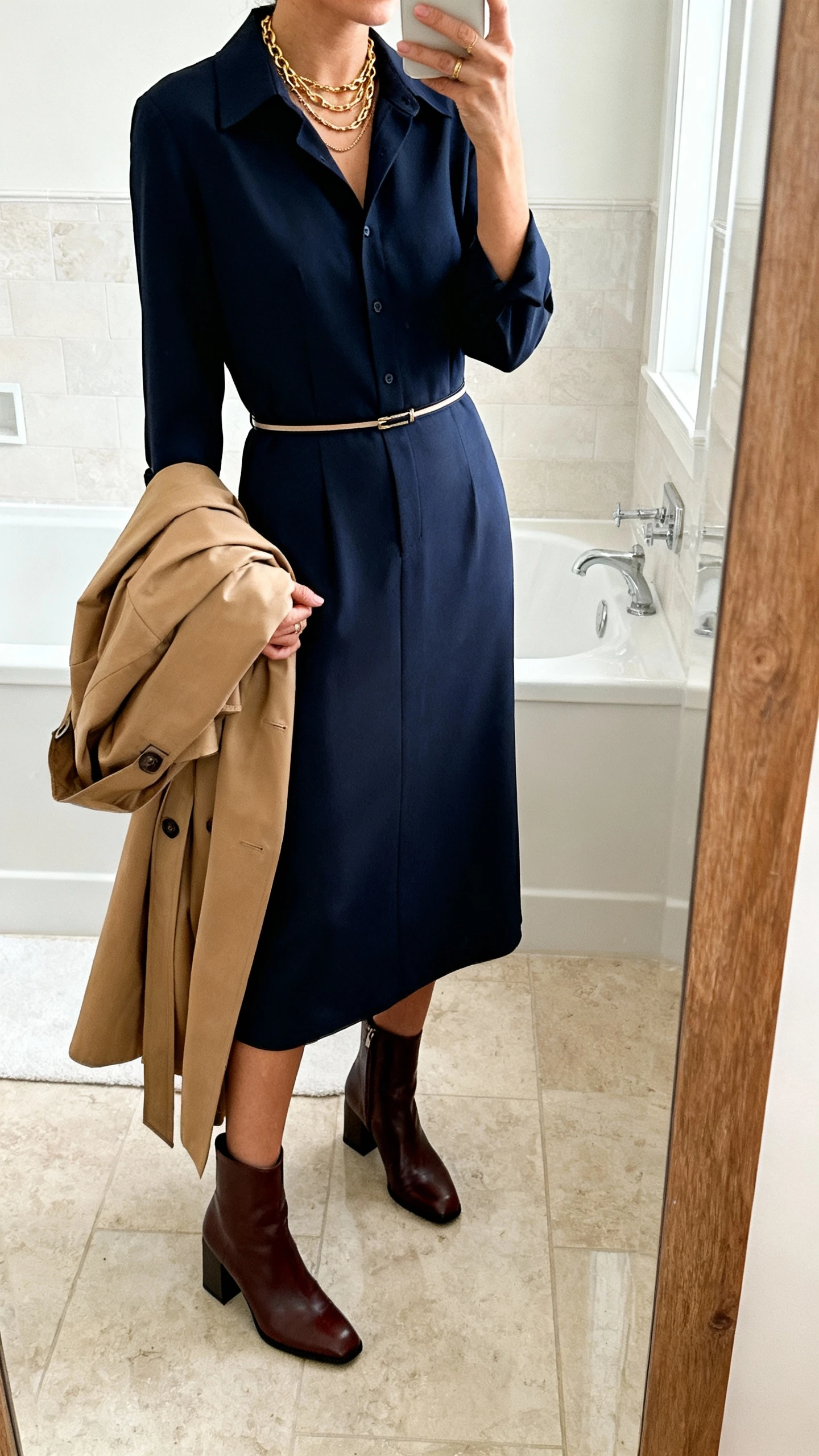 iPhone mirror selfie of a woman in a navy tailored midi shirt dress with a thin waist belt, leather ankle boots with a comfortable block heel, layered gold necklaces, and a lightweight camel trench draped over her arm, face not visible, bathroom mirror, soft indoor lighting, iPhone photo quality.