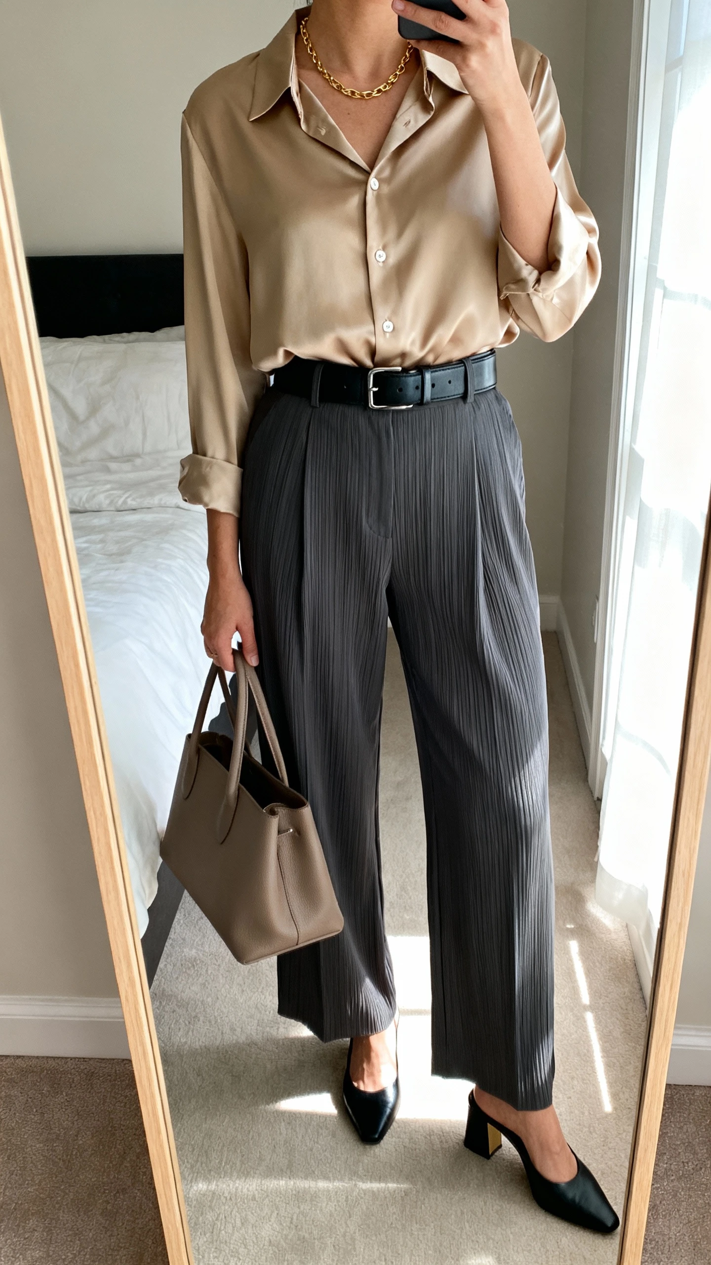 iPhone mirror selfie of a woman in a silk-meet-structure outfit: sand silk button-up blouse tucked into charcoal tailored pleated trousers, sleek black leather belt, black heeled mules, taupe structured tote, thin gold chain necklace, face not visible, cropped at shoulders, bedroom mirror with natural window light, iPhone photo quality