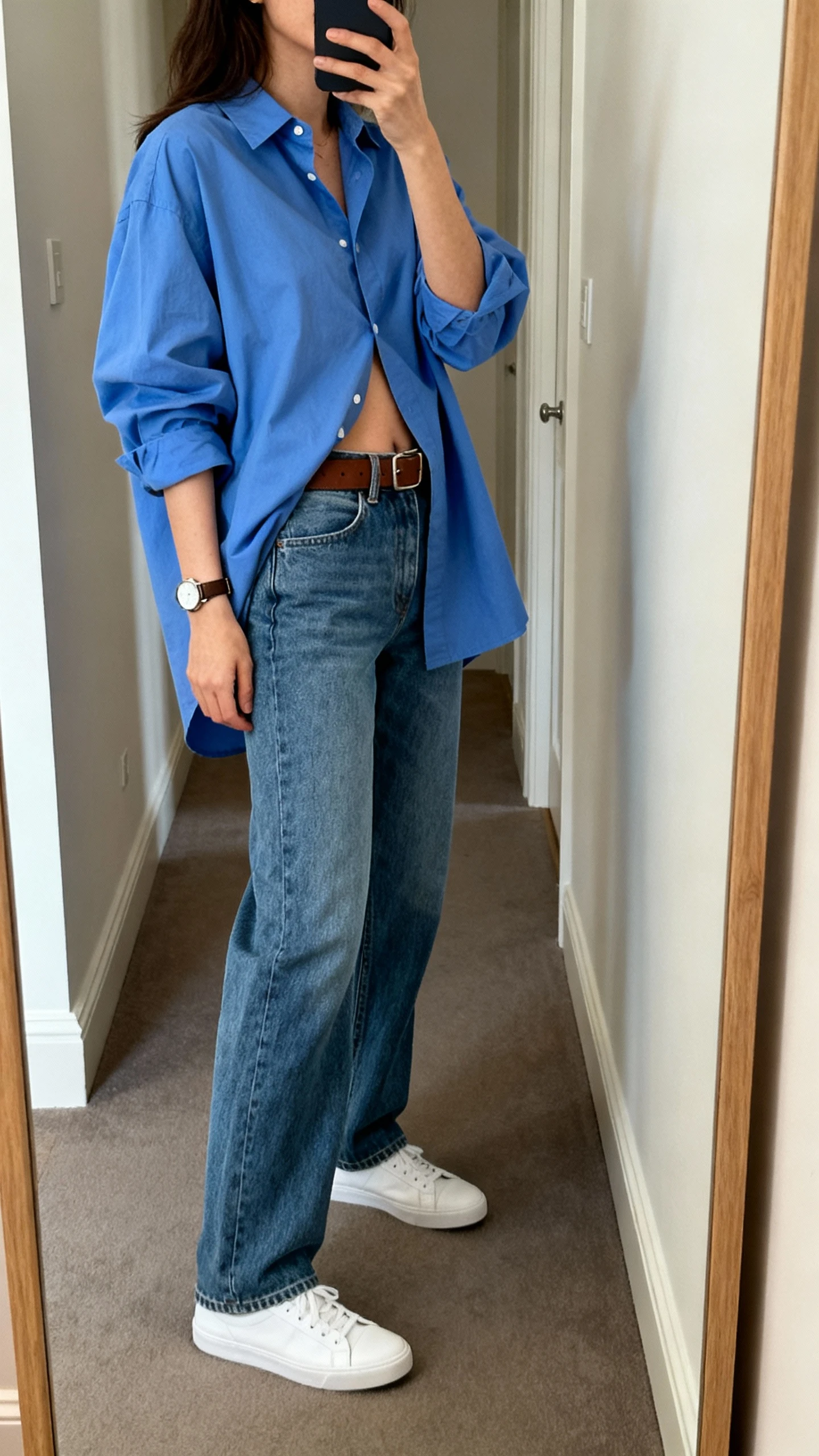 iPhone mirror selfie of a woman in relaxed straight-leg denim jeans with an oversized blue Oxford shirt half-tucked, classic leather belt, white low-top sneakers, simple watch, face not visible, side angle in a hallway full-length mirror, soft indoor lighting, iPhone photo quality