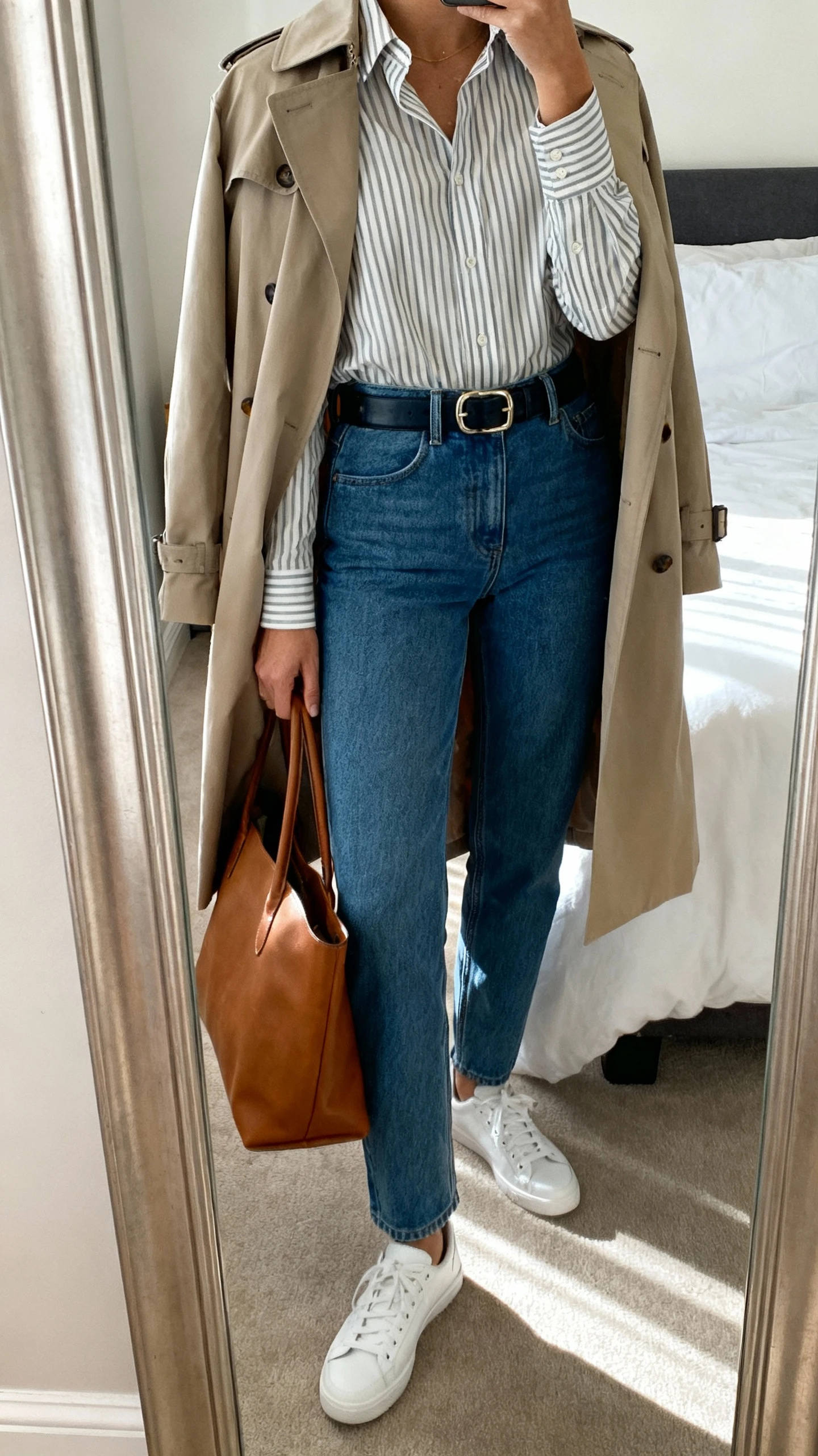 iPhone mirror selfie of a woman in straight-leg blue jeans, a tucked-in striped button-up shirt, polished belt, clean white sneakers, and a trench-style lightweight coat draped over shoulders with a leather tote, face not visible, cropped at shoulders, bedroom mirror, natural daylight, iPhone photo quality.