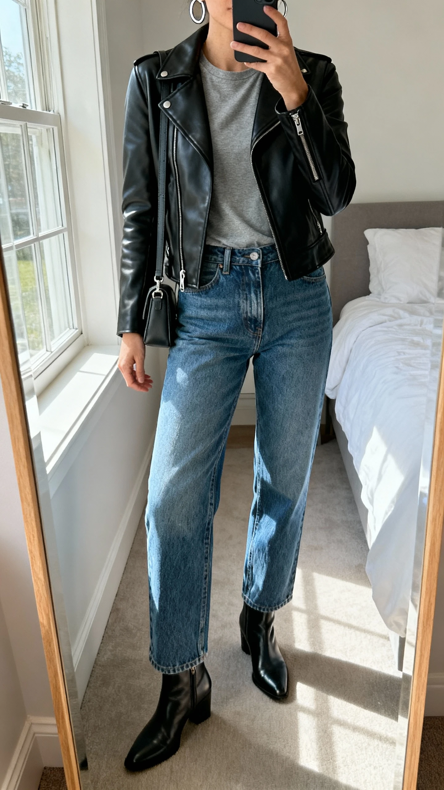 iPhone mirror selfie of a woman in straight-leg vintage blue jeans, a fitted black leather jacket over a simple grey tee, sleek ankle boots, a small shoulder bag, and silver hoops, face not visible, cropped at shoulders, bedroom mirror with natural window light, iPhone photo quality.