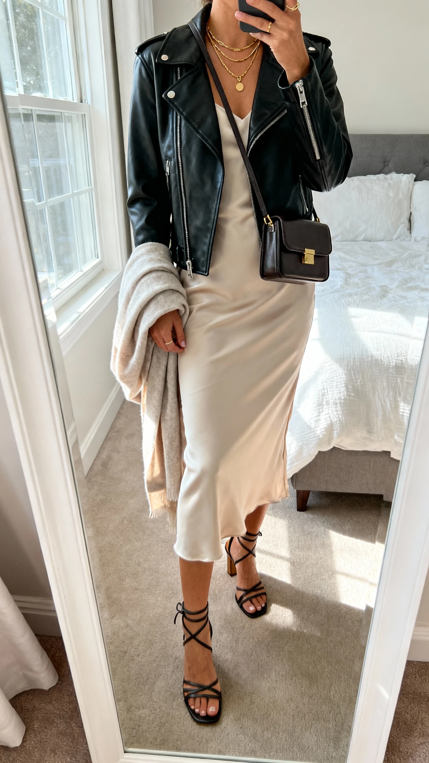 iPhone mirror selfie of a woman wearing a bias-cut silk midi slip dress with a black leather moto jacket, strappy heeled sandals, delicate gold layered necklaces, a structured mini shoulder bag, and a soft shawl draped over her arm; face not visible, cropped at neck, bedroom mirror, natural window light, iPhone photo quality.