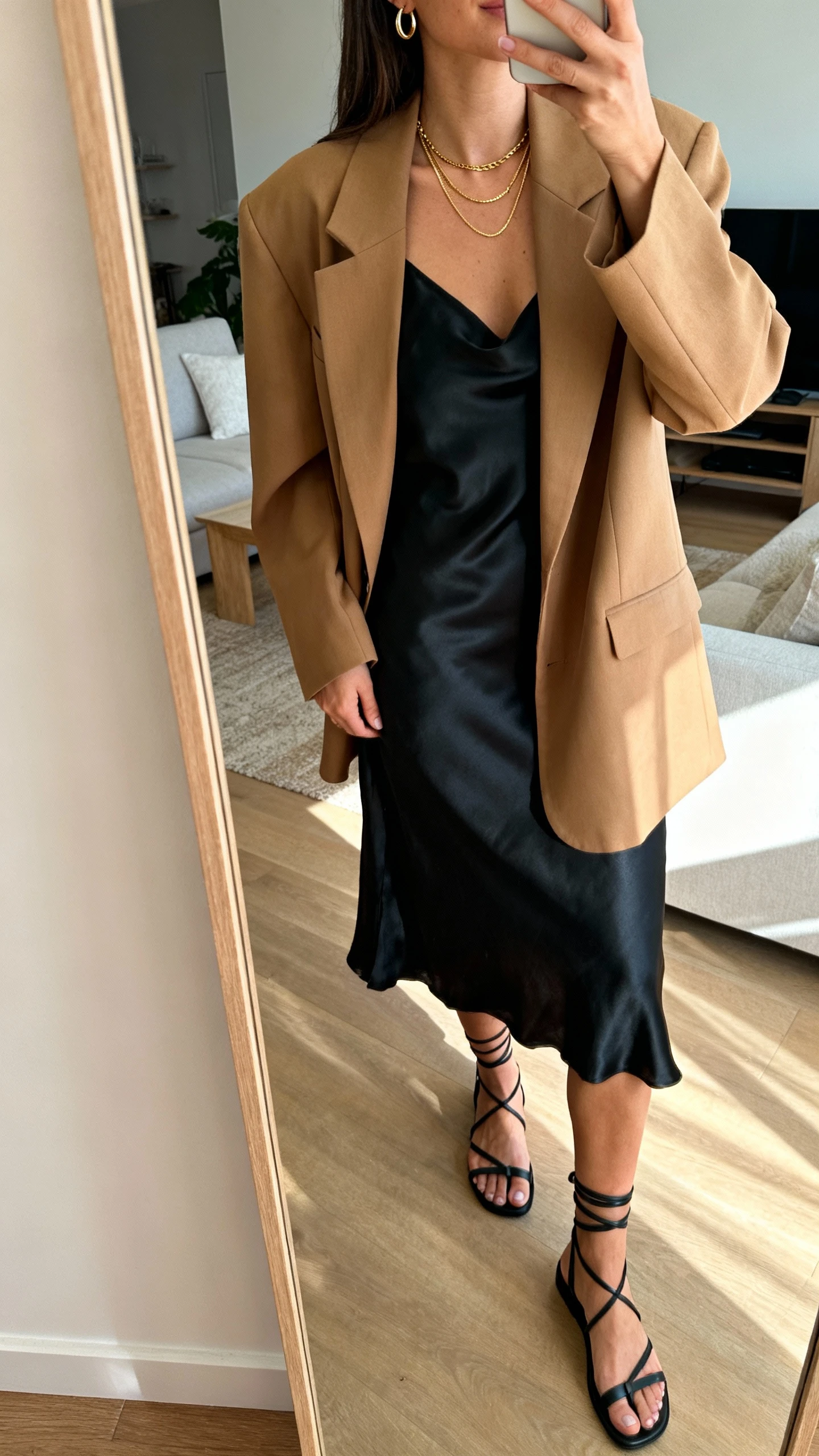 iPhone mirror selfie of a woman wearing a black satin slip dress under an oversized camel blazer, delicate layered gold necklaces, thin hoop earrings, and strappy black sandals; face not visible, cropped at neck, living-room full-length mirror with soft afternoon natural light, iPhone photo quality.