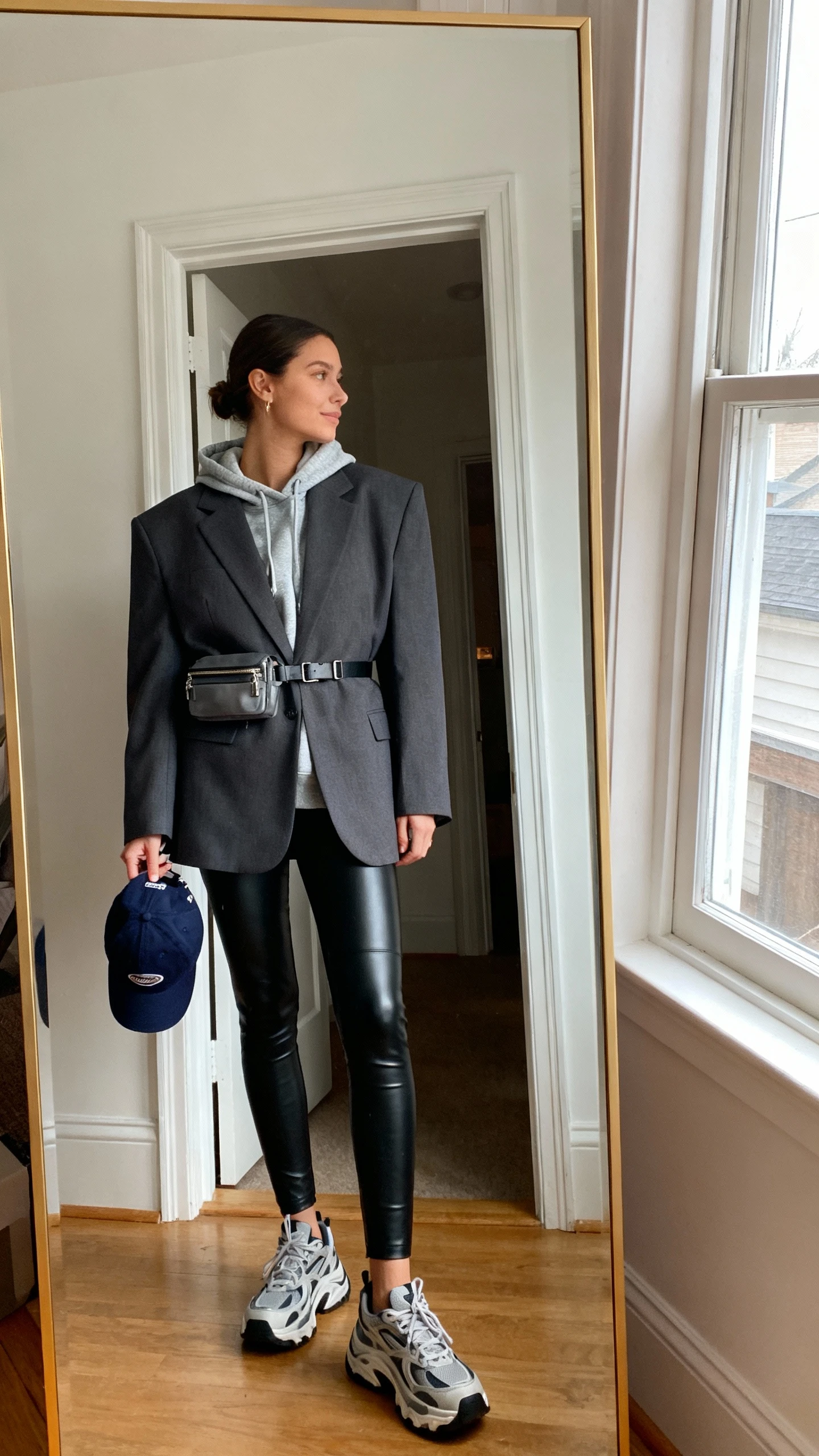 iPhone mirror selfie of a woman wearing a boxy blazer layered over a hoodie with leather leggings, chunky sneakers, a baseball cap in hand, and a small belt bag, face not visible, side angle, entryway full-length mirror, natural window light, iPhone photo quality.