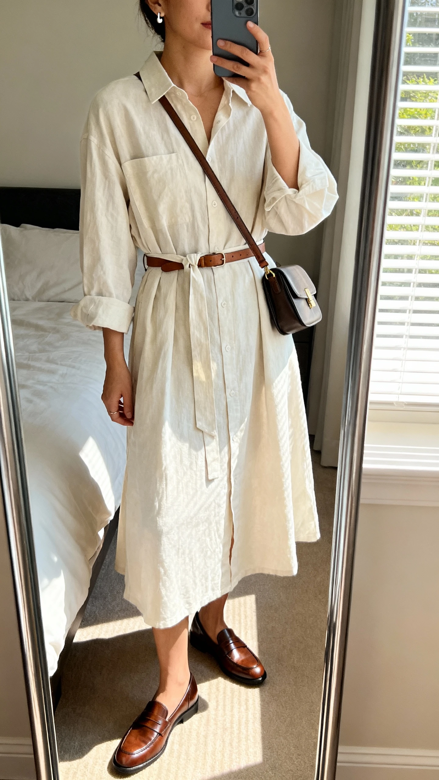 iPhone mirror selfie of a woman wearing a breezy cotton shirt dress with a waist tie, sleek leather loafers, minimalist stud earrings, a slim leather belt, and a structured crossbody bag, face not visible, cropped at neck, bedroom mirror with natural window light, iPhone photo quality.