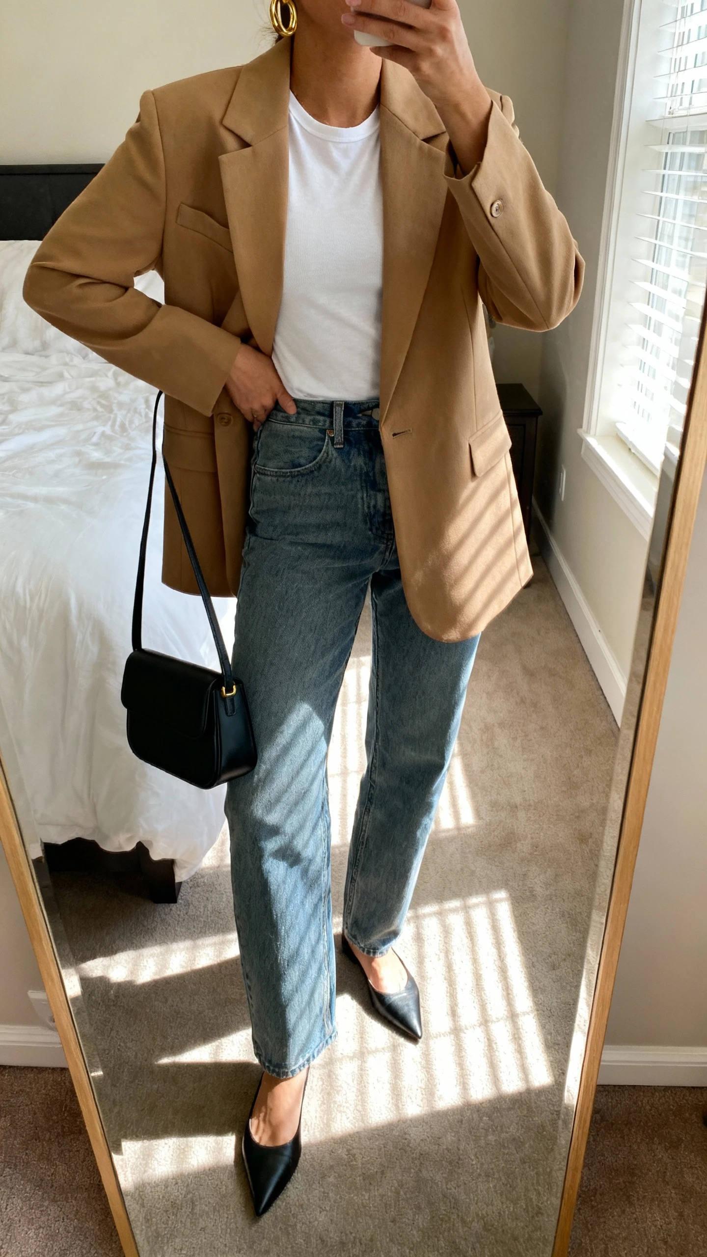 iPhone mirror selfie of a woman wearing a camel tailored blazer over a white fitted tee, relaxed straight-leg medium-wash jeans, pointed-toe flats, a structured black mini tote, and gold hoop earrings, face not visible, cropped at neck, bedroom mirror with morning window light, iPhone photo quality.