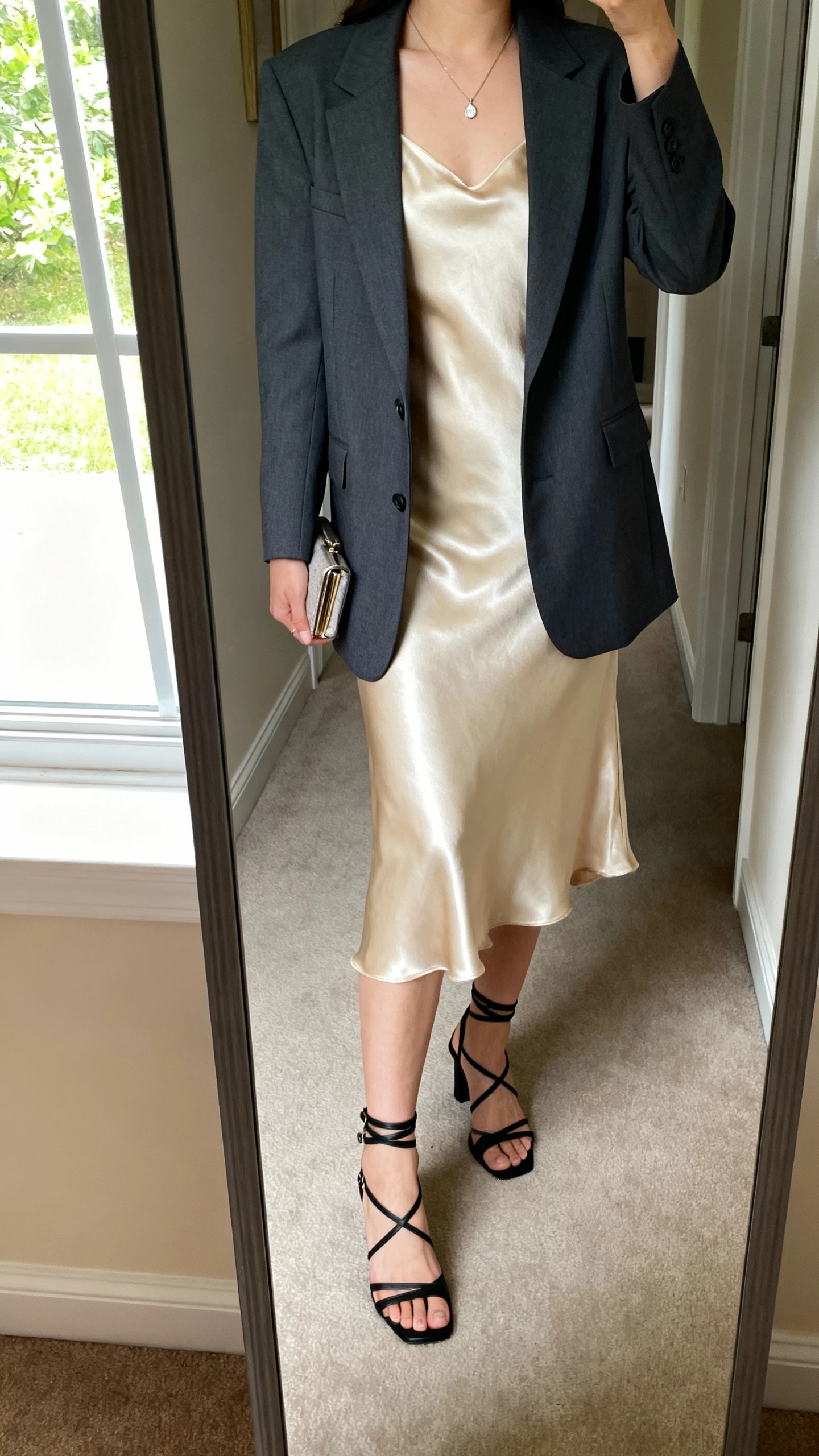iPhone mirror selfie of a woman wearing a champagne satin slip dress layered with a tailored charcoal blazer, strappy black heels, delicate pendant necklace, and a petite clutch, face not visible, cropped at neck, full-length mirror in hallway, morning light from window, iPhone photo quality.