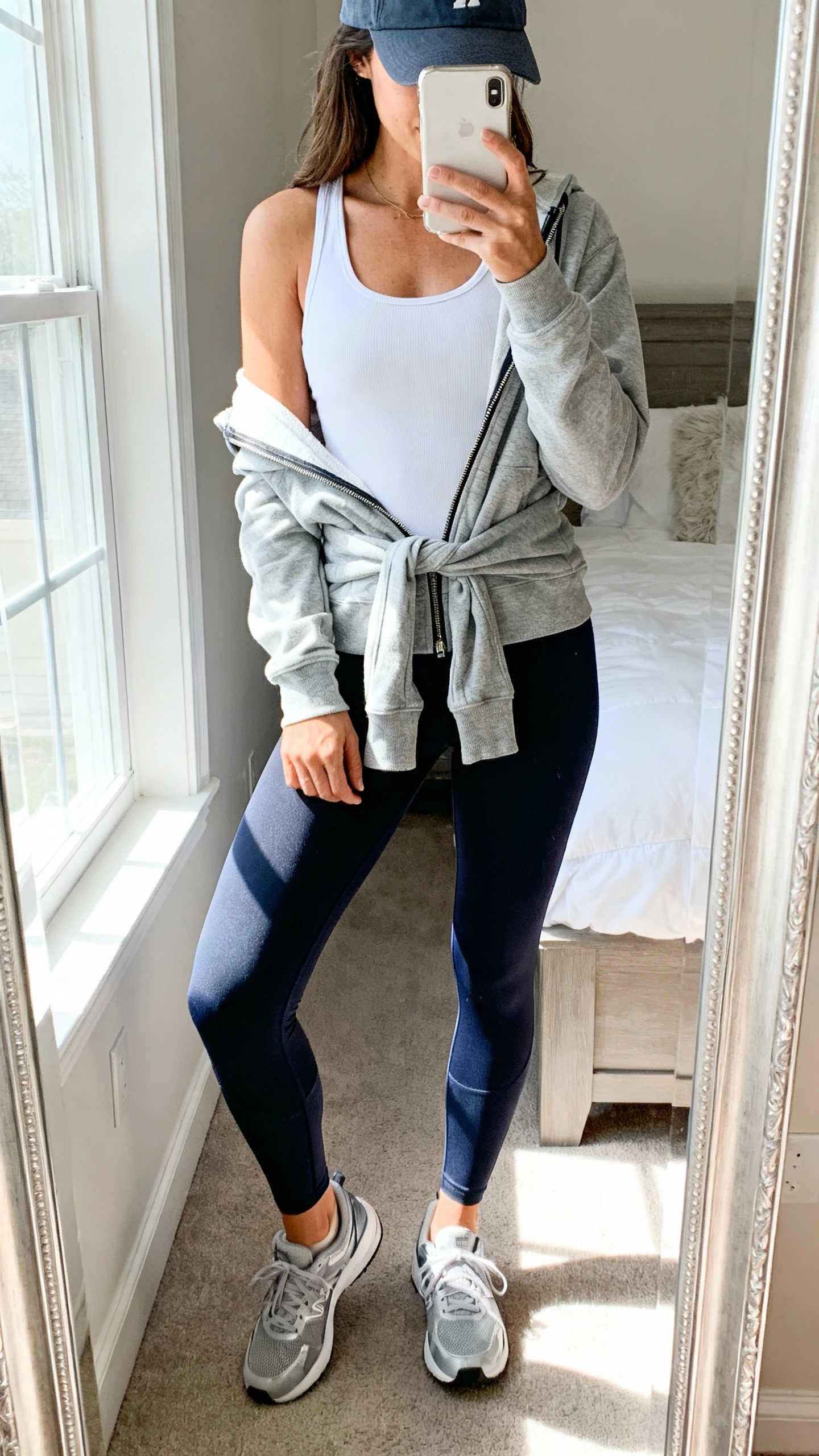 iPhone mirror selfie of a woman wearing a chic athleisure mix: performance leggings, breathable tank, lightweight zip-up hoodie tied at waist, sleek trainers and a baseball cap, face not visible, cropped at neck, bedroom mirror, bright window light, iPhone photo quality.