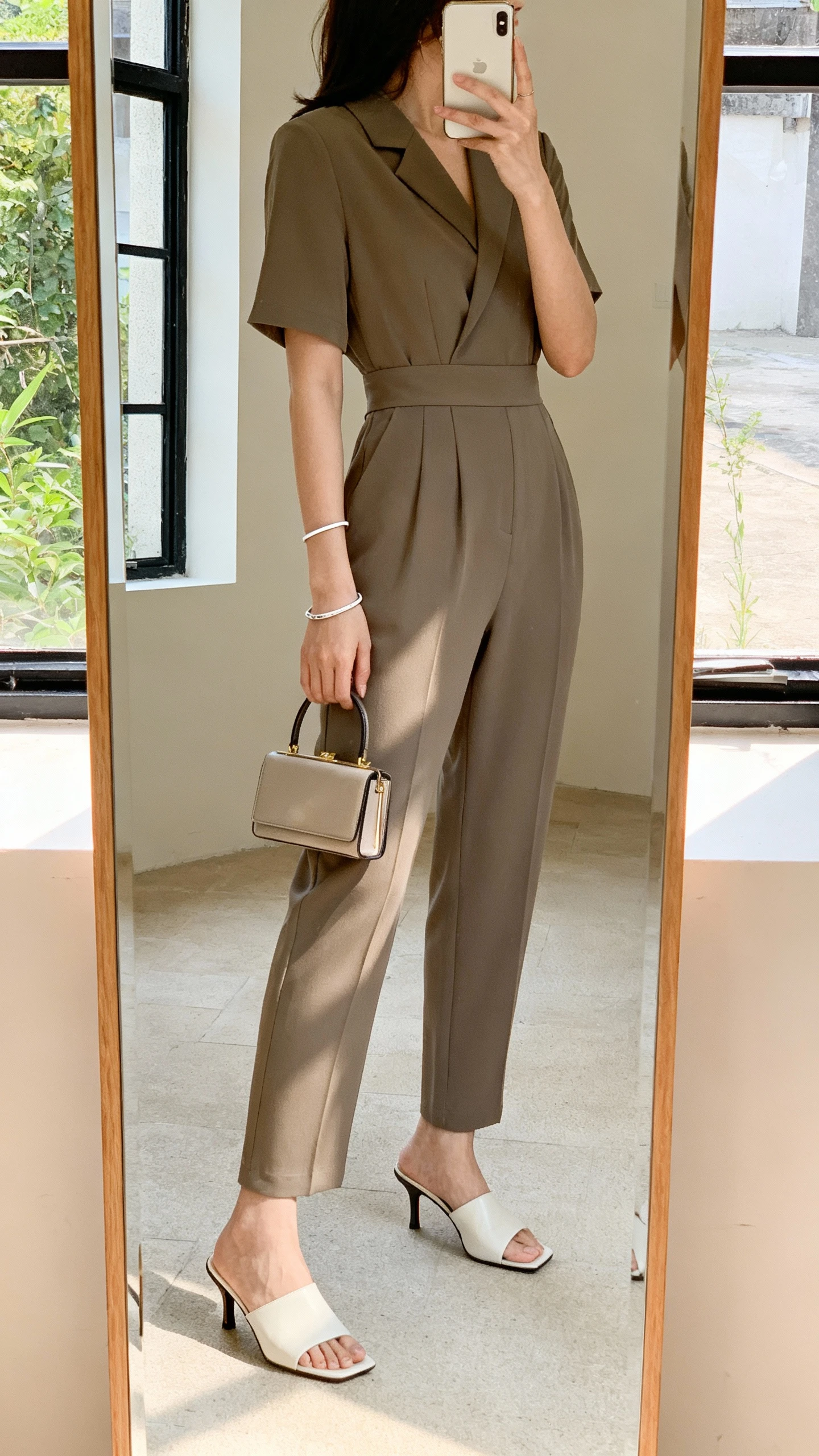 iPhone mirror selfie of a woman wearing a chic tailored jumpsuit with a defined waist, heeled mules, minimalist bracelet, and a compact handbag, face not visible, side profile in full-length mirror, soft natural daylight from window, iPhone photo quality.
