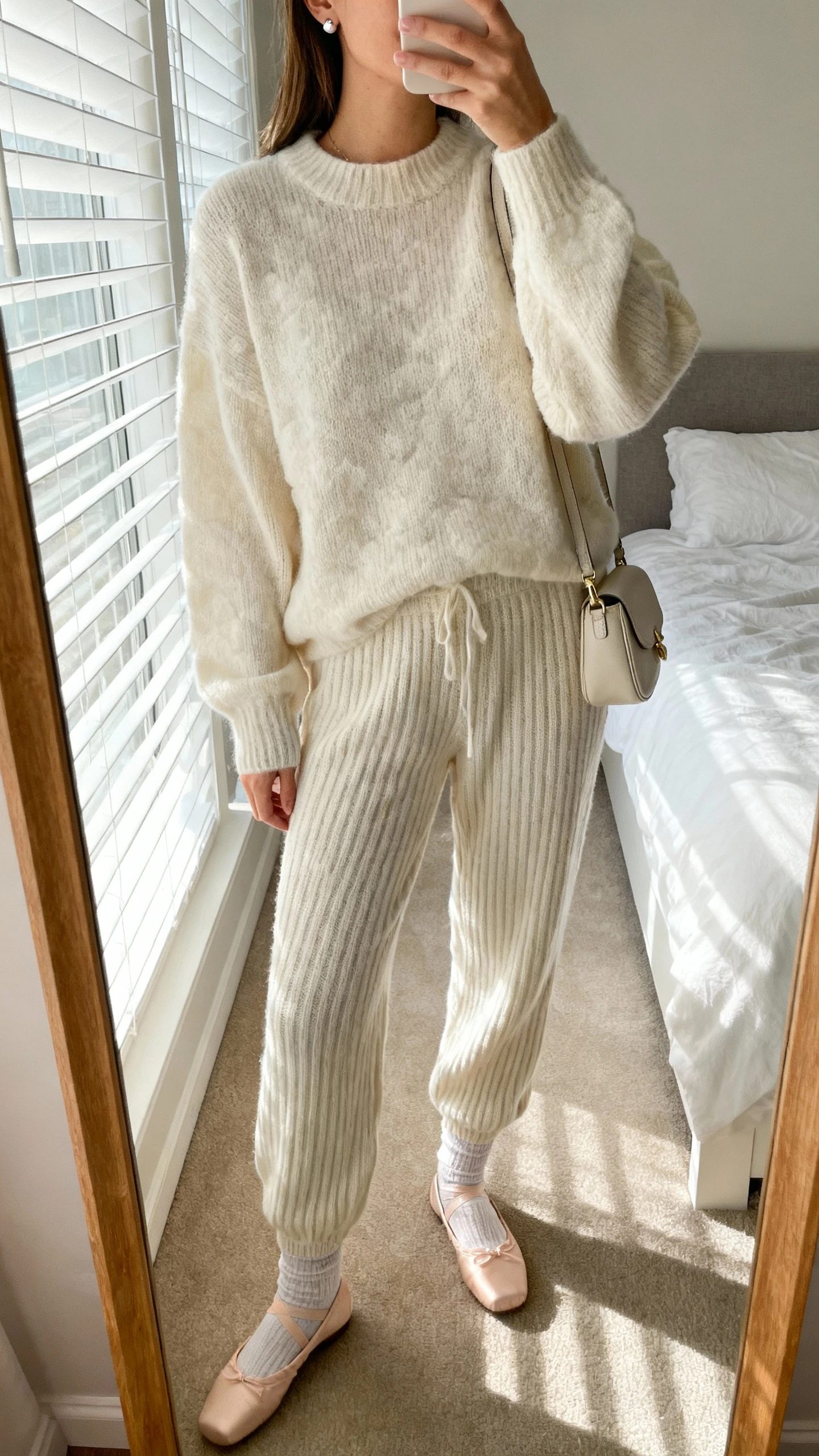 iPhone mirror selfie of a woman wearing a cloud-soft matching knit set (ribbed sweater and knit lounge pants) with ballet flats, delicate socks, small shoulder bag, simple stud earrings, face not visible, cropped at neck, bedroom mirror, soft morning window light, iPhone photo quality
