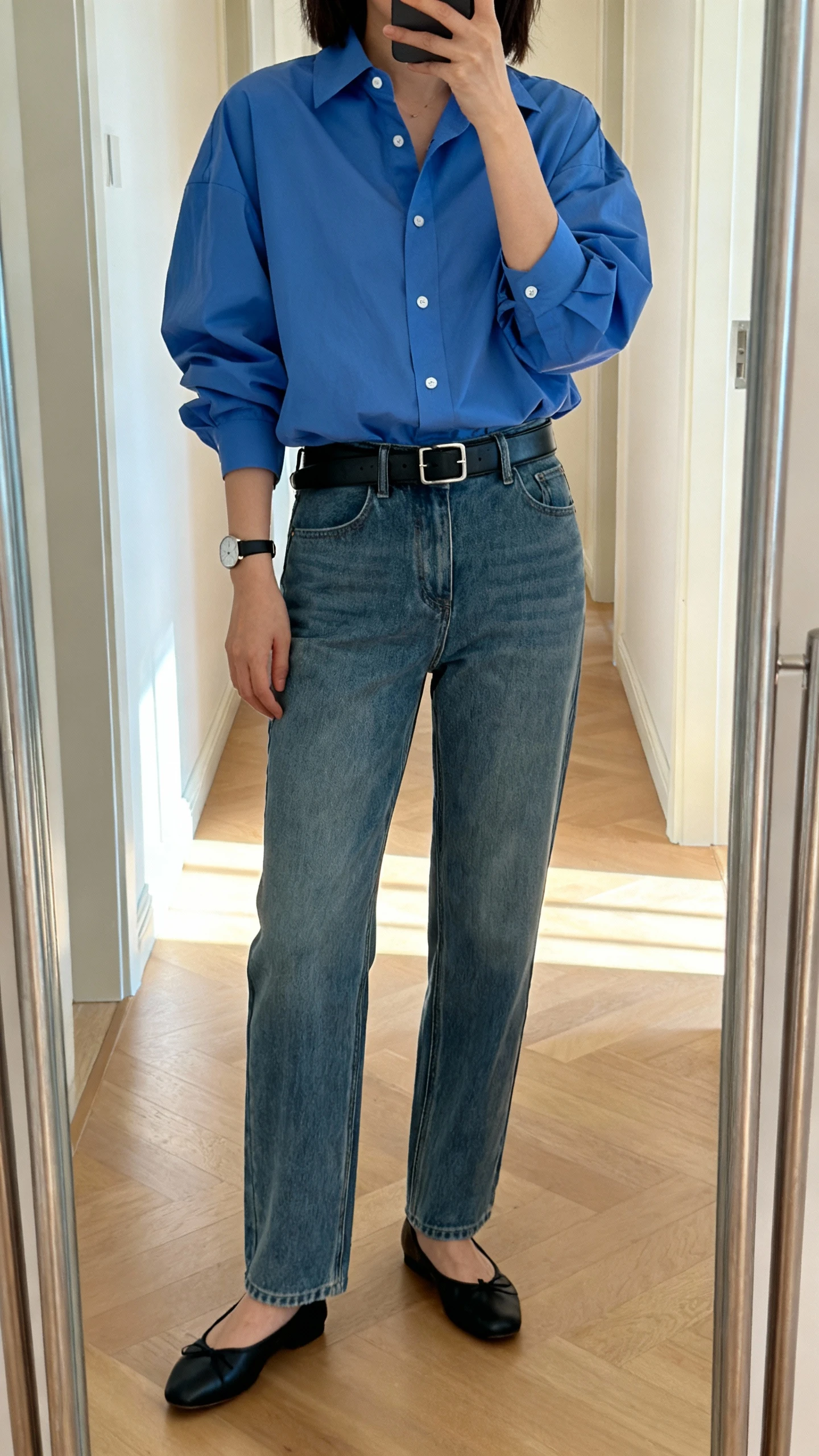 iPhone mirror selfie of a woman wearing a crisp oversized blue button-down tucked into straight-leg medium-wash jeans, black leather belt, classic black ballet flats, and a minimalist watch; face not visible, cropped at shoulders, full-length hallway mirror with natural daylight, iPhone photo quality.
