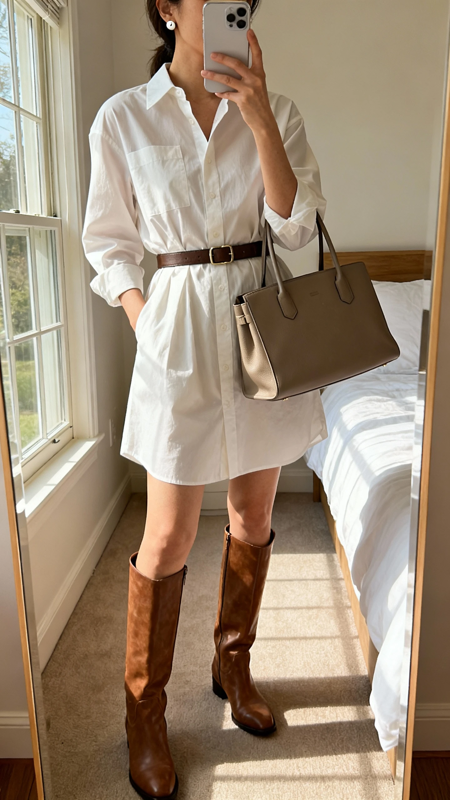 iPhone mirror selfie of a woman wearing a crisp shirt dress cinched with a waist belt, knee-high boots, structured tote, and simple stud earrings, face not visible, cropped at shoulders, bedroom mirror with afternoon window light, iPhone photo quality.