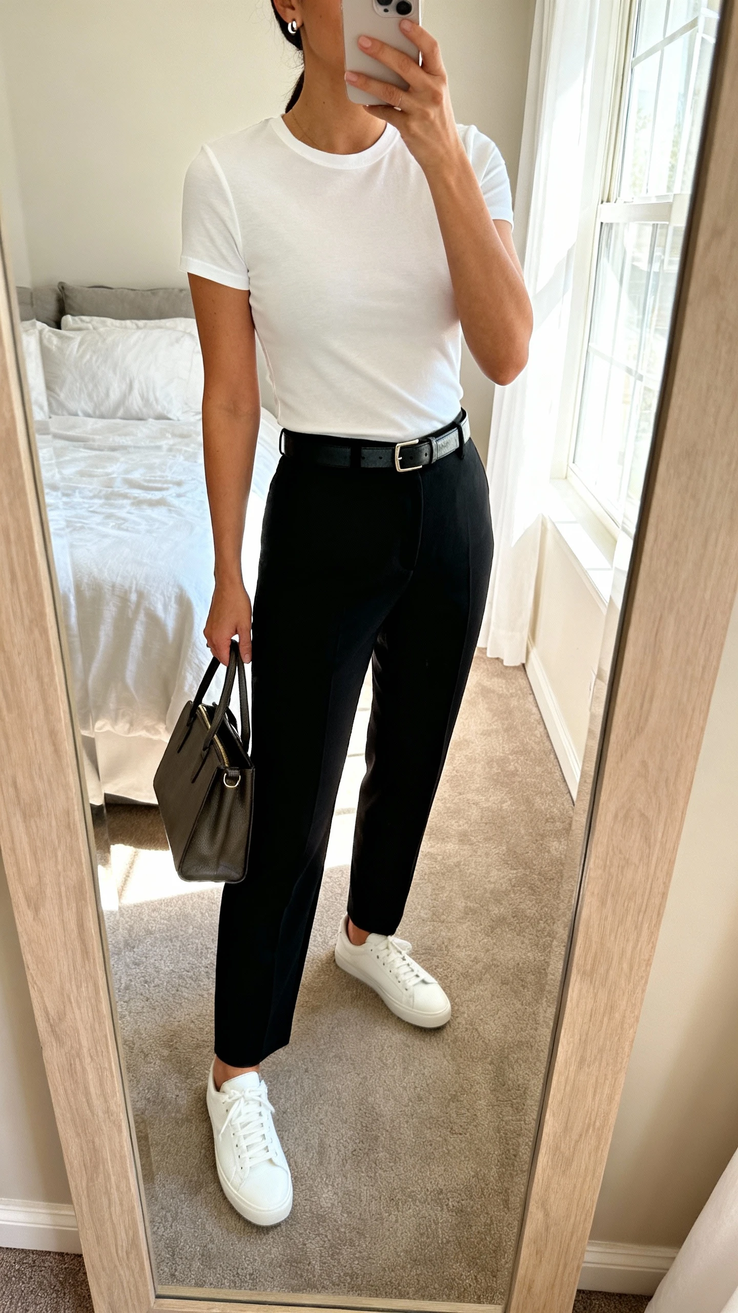 iPhone mirror selfie of a woman wearing a crisp white fitted tee tucked into tailored black trousers with white low-top sneakers, a slim leather belt, minimalist stud earrings, and a structured tote, face not visible, cropped at neck, bedroom mirror, soft morning window light, iPhone photo quality.