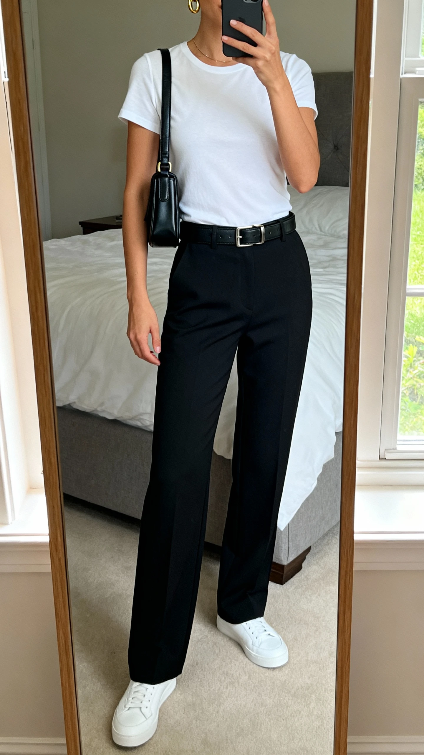 iPhone mirror selfie of a woman wearing a crisp white fitted tee tucked into tailored black trousers, sleek leather belt, minimalist white sneakers, dainty gold hoops, structured black crossbody, face not visible, cropped at neck, bedroom mirror, natural window light, iPhone photo quality.