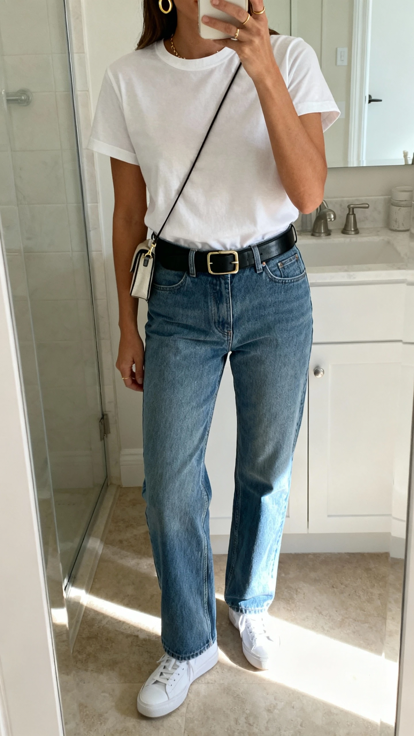 iPhone mirror selfie of a woman wearing a crisp white elevated tee tucked into straight-leg medium-wash jeans, black leather belt, classic white sneakers, delicate gold hoops, and a small crossbody bag, face not visible, bathroom mirror, morning light, iPhone photo quality.