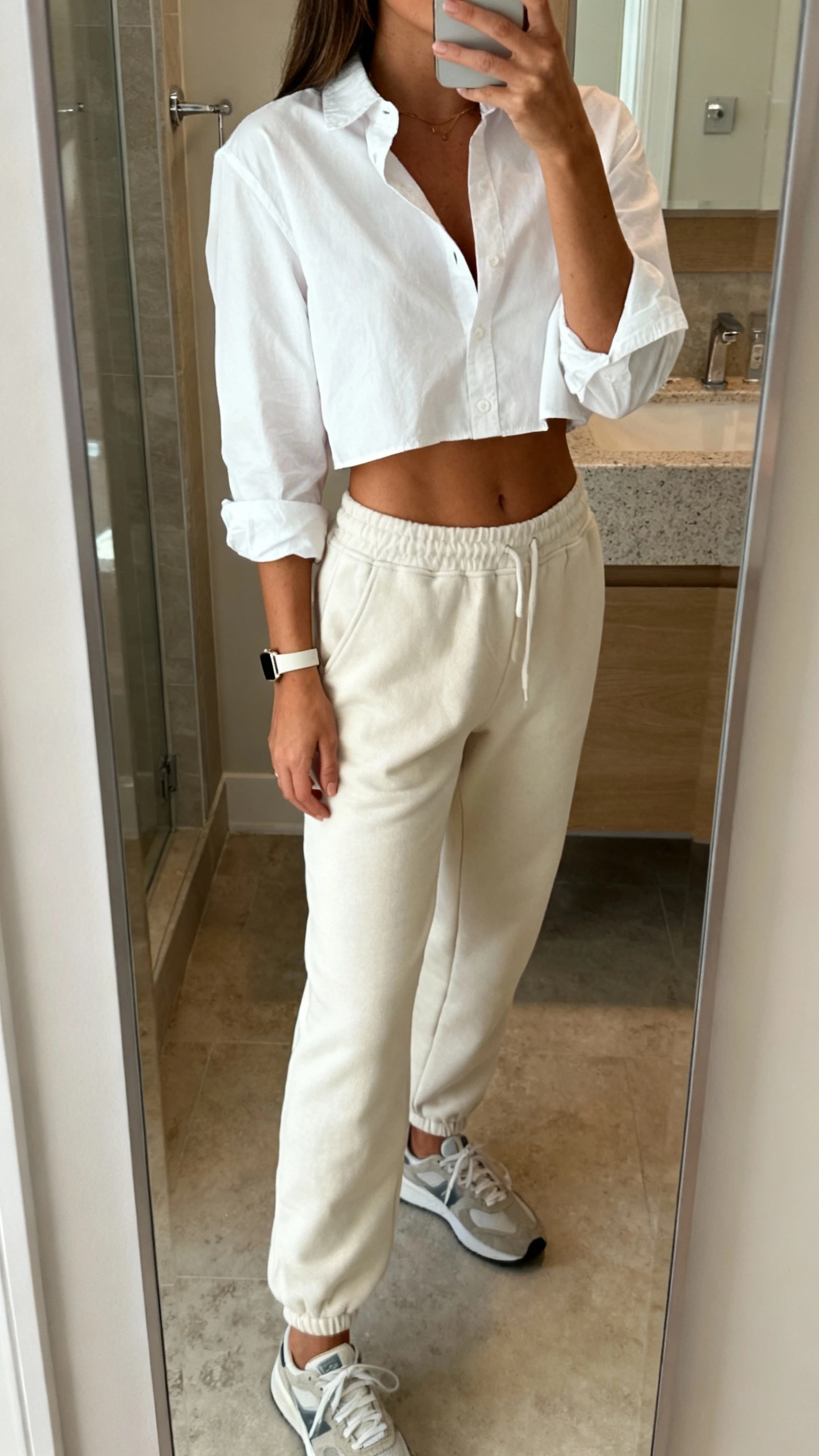 iPhone mirror selfie of a woman wearing a crisp white button-up shirt half-tucked into soft joggers, with low-profile sneakers and a slim watch, face not visible, bathroom full-length mirror, natural indoor lighting, iPhone photo quality.