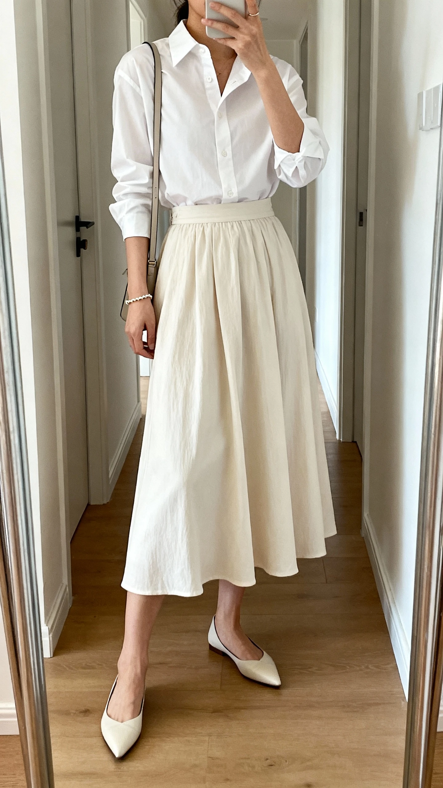iPhone mirror selfie of a woman wearing a crisp white button-down shirt tucked into a flowy midi skirt, pointed flats, delicate bracelet, and a slim shoulder bag, face not visible, cropped at shoulders, full-length hallway mirror, natural indoor light, iPhone photo quality