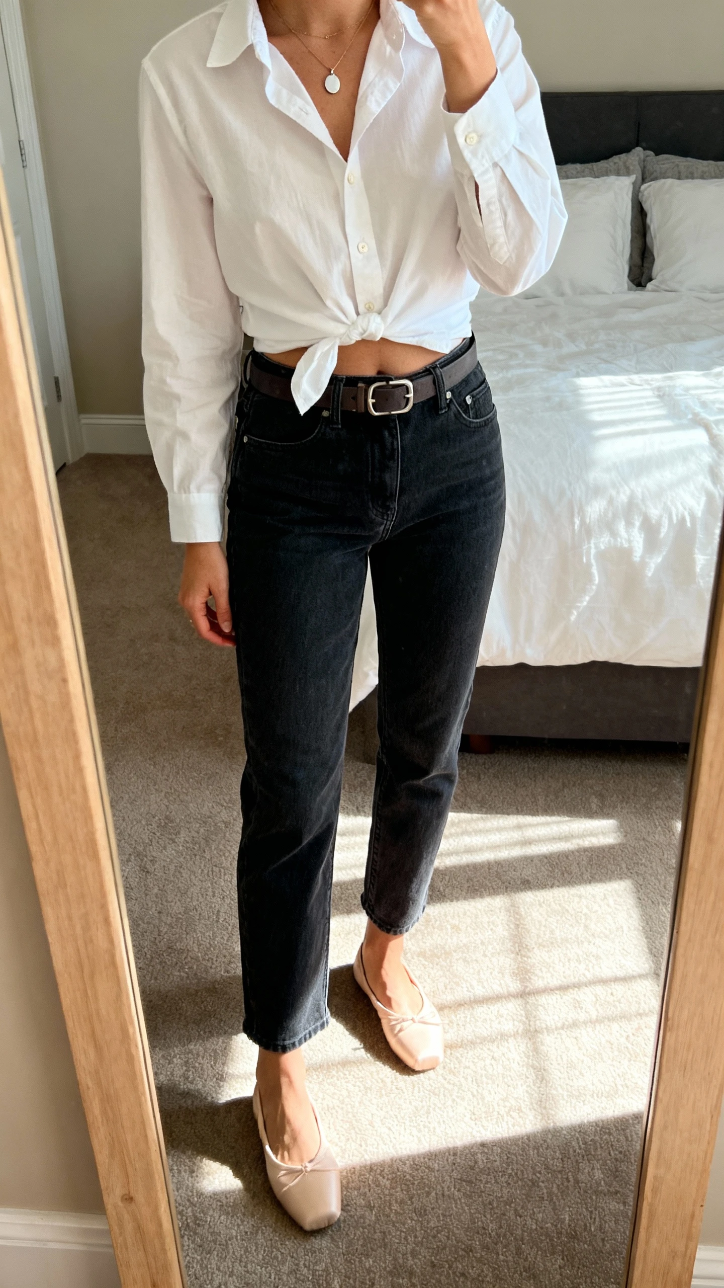 iPhone mirror selfie of a woman wearing a crisp white button-down half-tucked into dark wash straight-leg jeans, belt, ballet flats, and a simple pendant necklace, face not visible, cropped at shoulders, bedroom mirror, natural morning light, iPhone photo.