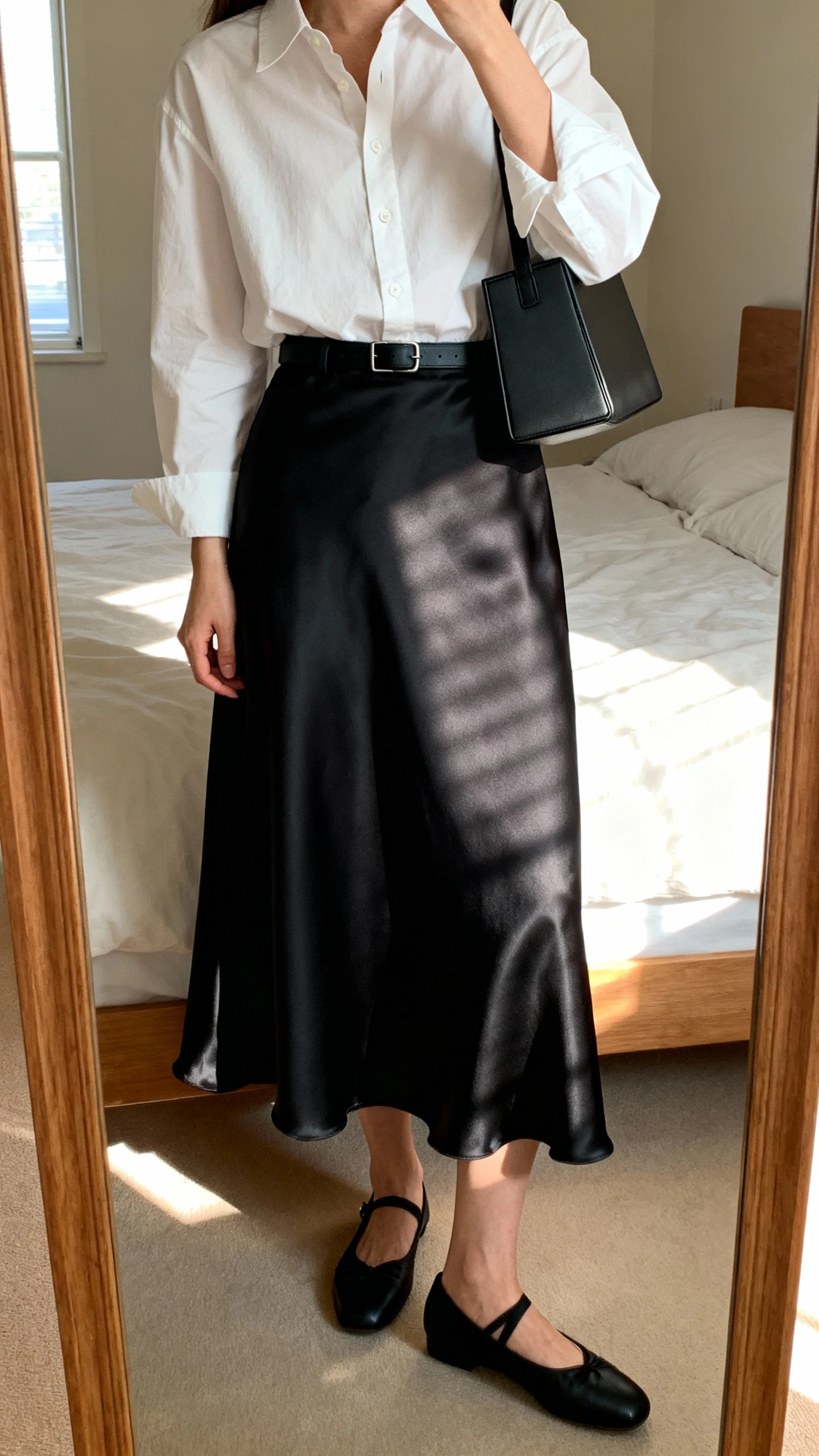 iPhone mirror selfie of a woman wearing a crisp white cotton-poplin button-down tucked into a flowy black satin midi skirt, refined black ballet flats, a minimal black belt, and a boxy shoulder bag, face not visible, cropped at shoulders, bedroom mirror with morning window light, iPhone photo quality.