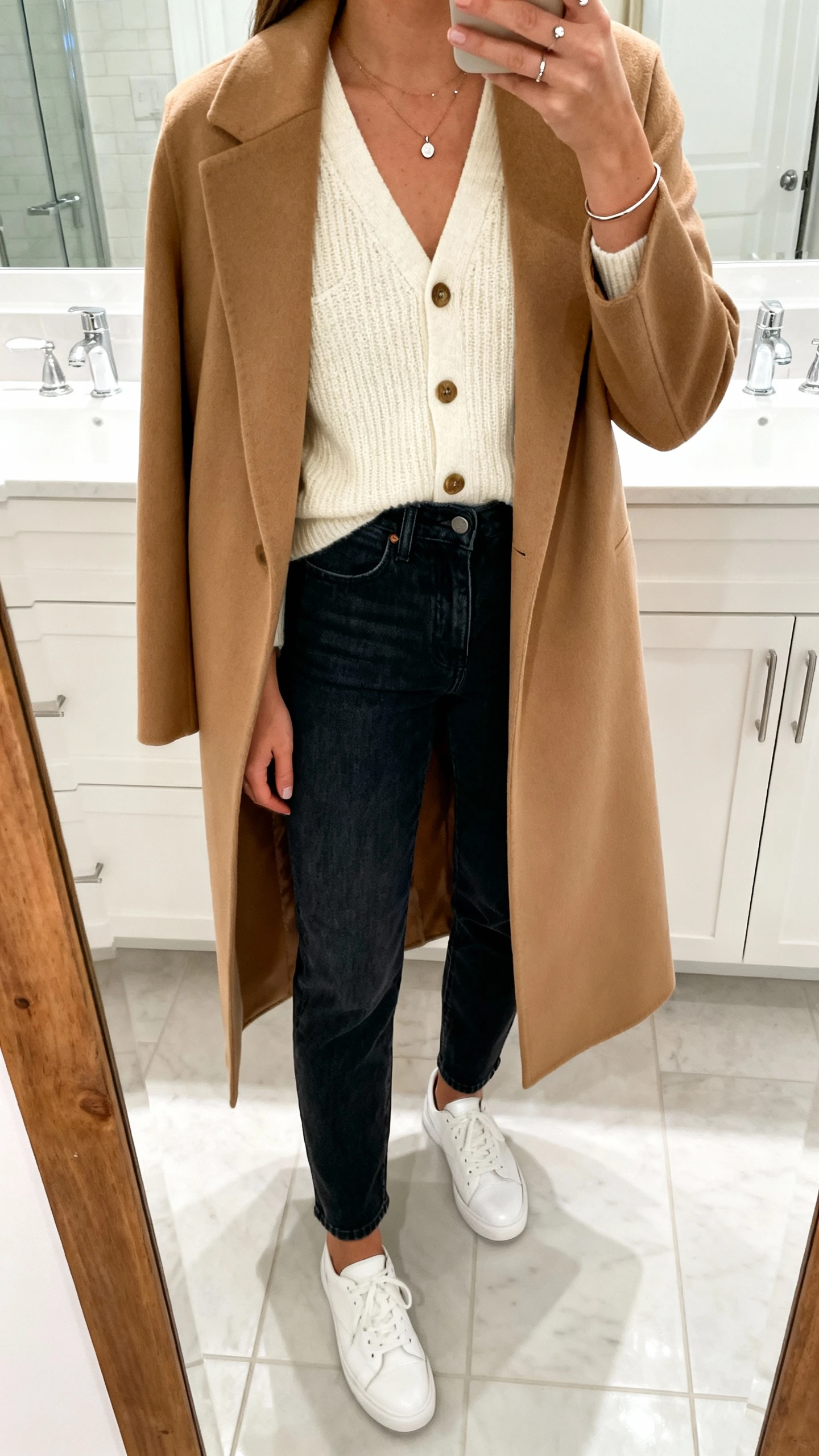 iPhone mirror selfie of a woman wearing a fine-knit cream cardigan buttoned as a top, high-rise dark-wash straight jeans, minimal white leather sneakers, delicate pendant necklace, studs, slim bangle, and a tailored camel longline coat; face not visible, cropped at neck, bathroom mirror with bright indoor lighting, iPhone photo quality.