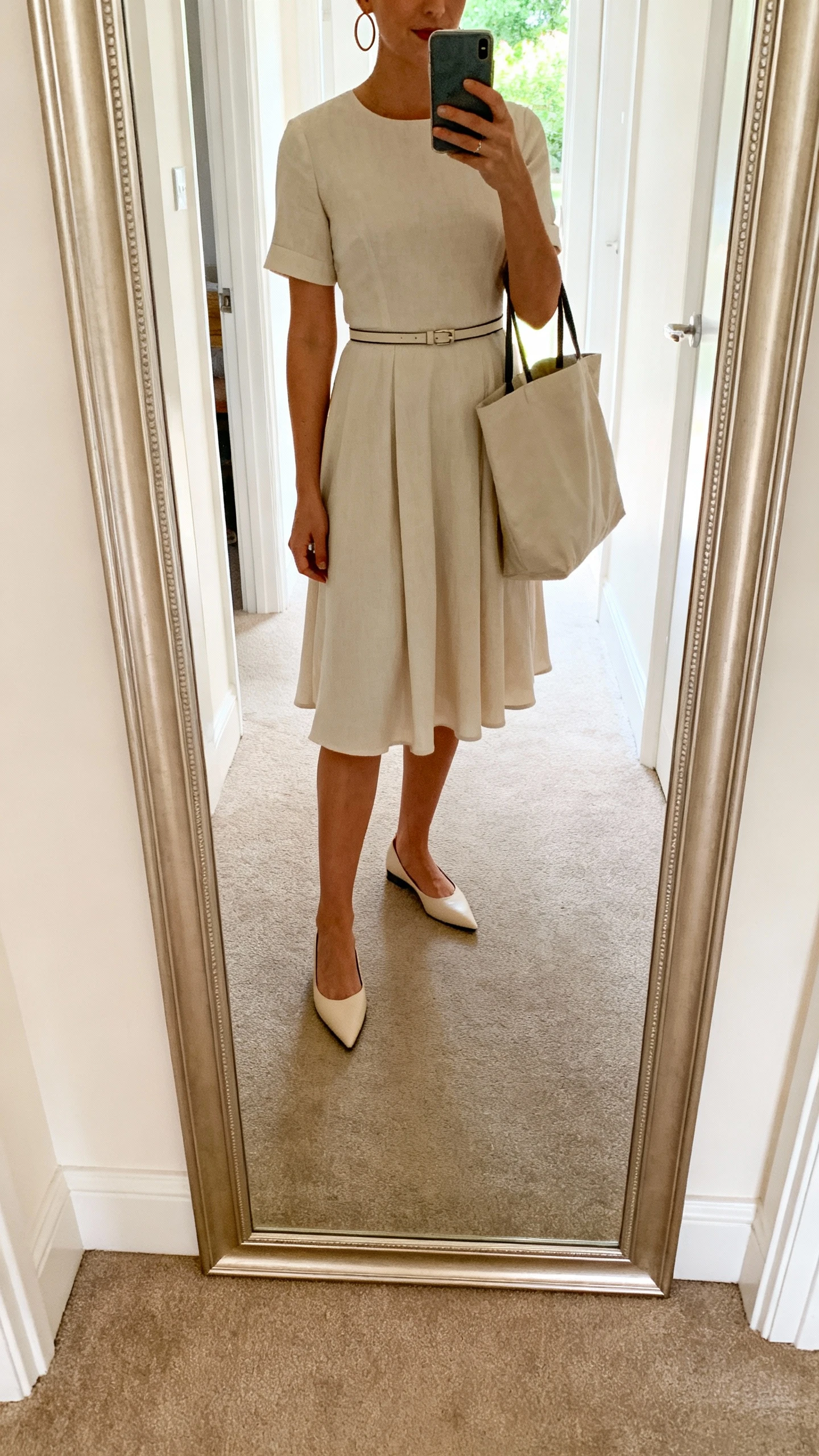 iPhone mirror selfie of a woman wearing a fit-and-flare day dress, pointed-toe flats, thin waist belt, simple hoop earrings, and a tote bag, face not visible, cropped at shoulders, full-length mirror in hallway, bright indoor daylight, iPhone photo quality.