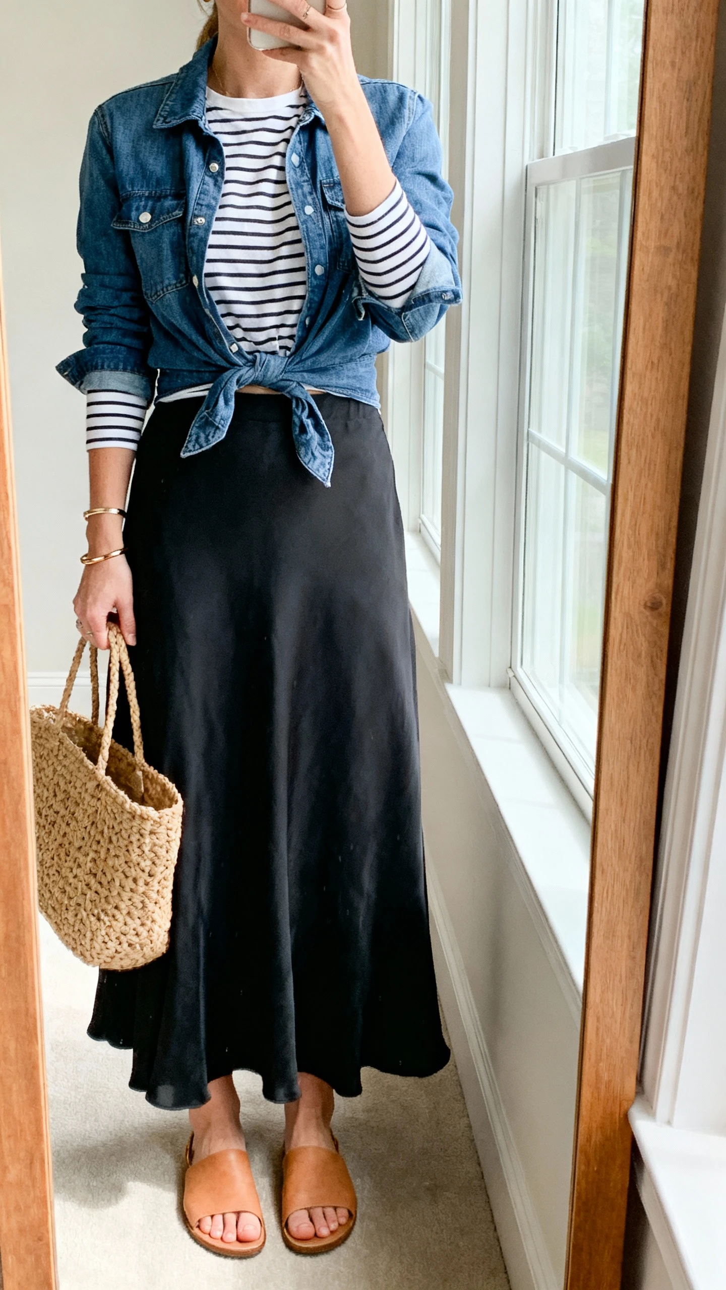 iPhone mirror selfie of a woman wearing a flowy black maxi skirt, striped long-sleeve tee half-tucked, denim jacket tied at the waist, tan flat sandals, woven tote, slim bracelets, face not visible, cropped at neck, full-length mirror by window, soft morning light, iPhone photo quality.