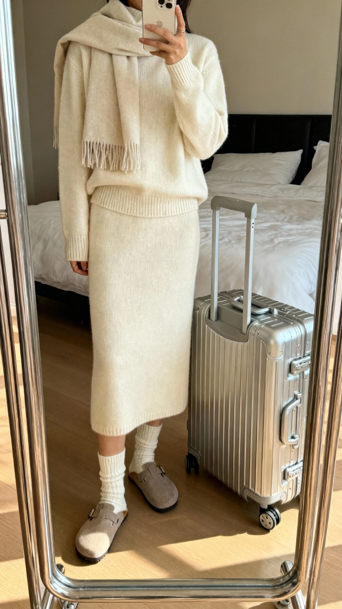 iPhone mirror selfie of a woman wearing a matching cashmere sweater set: crewneck sweater and knit midi skirt in cream, cozy socks with slip-on clogs, oversized scarf, sleek roller suitcase by her side, face not visible, cropped at shoulders, bedroom mirror, warm indoor lighting, iPhone photo quality.