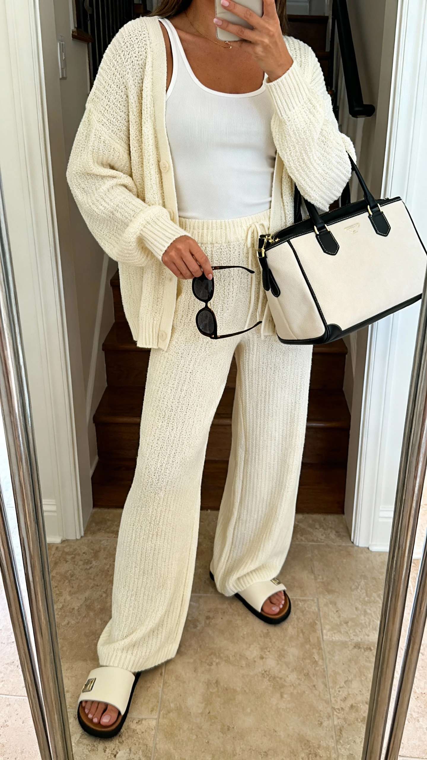 iPhone mirror selfie of a woman wearing a matching knit lounge set (cream cardigan and wide-leg pants) with a fitted tank, slide sandals, oversized sunglasses in hand, and a weekender bag, face not visible, full-length mirror by entryway, warm indoor lighting, iPhone photo quality.