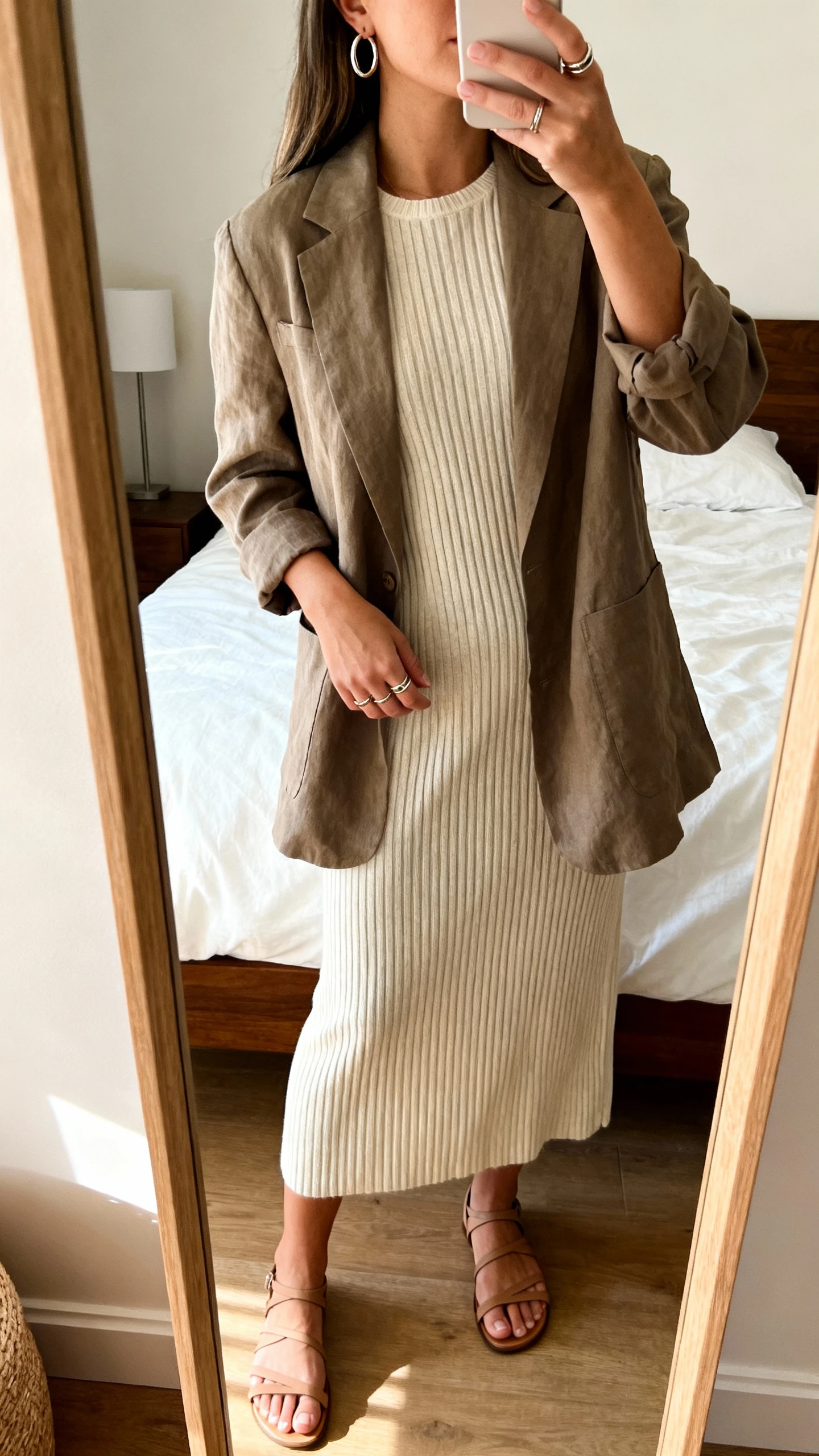 iPhone mirror selfie of a woman wearing a ribbed knit midi dress under a relaxed blazer, simple hoop earrings, stacked rings, and minimalist strappy sandals, face not visible, cropped at shoulders, bedroom mirror, warm indoor light, iPhone photo quality.